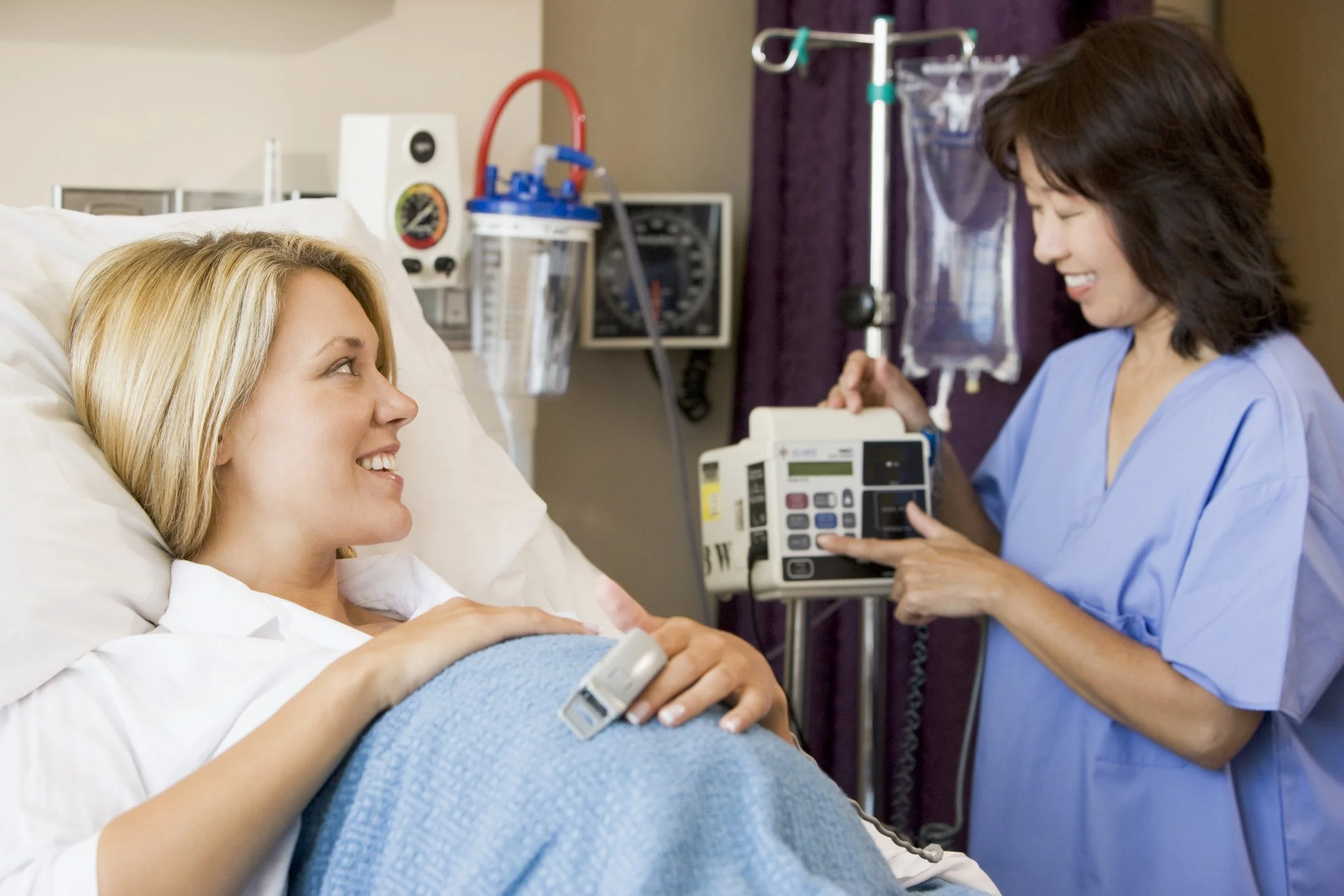 A smiling woman lying in a hospital bed talking to a nurse who is operating medical equipment.