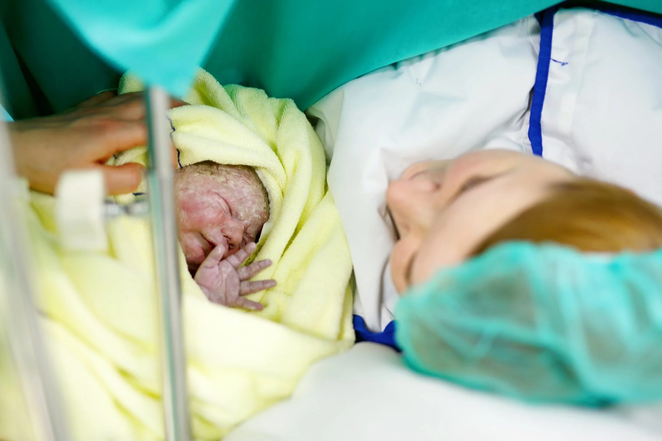 A newborn baby wrapped in a yellow towel next to a woman lying in a hospital bed, wearing a green hospital gown and cap, in a hospital delivery room.
