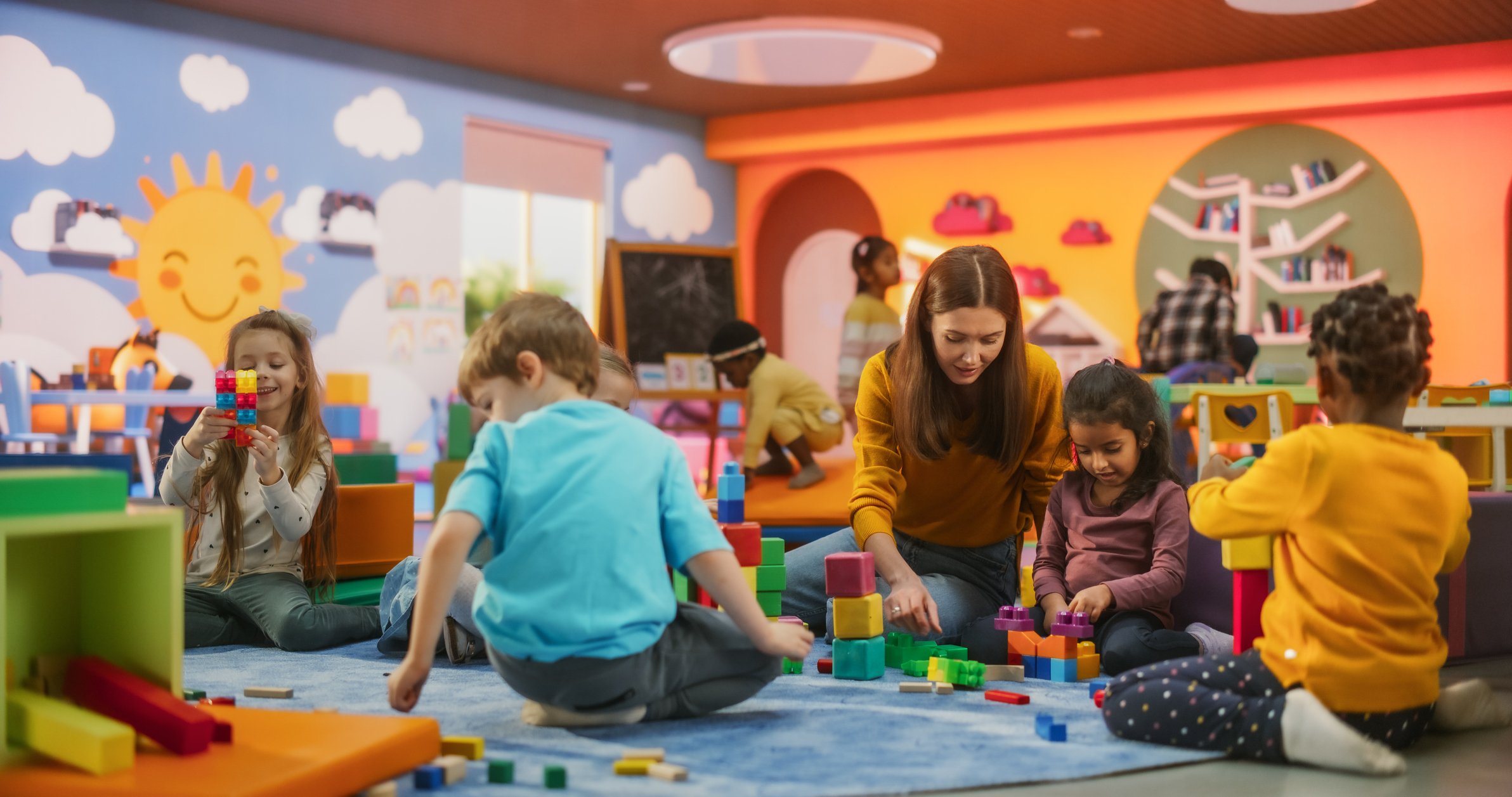 Children and a woman playing with building blocks in a colorful playroom.