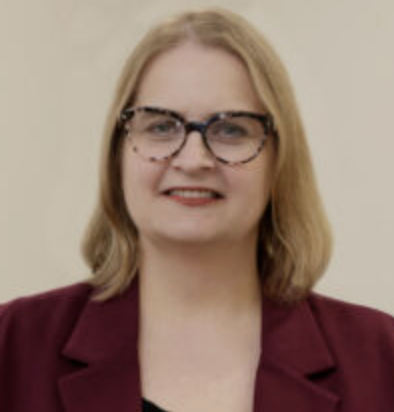 A woman with shoulder-length blonde hair, wearing glasses and a burgundy blazer, smiling in front of a plain light-colored background.
