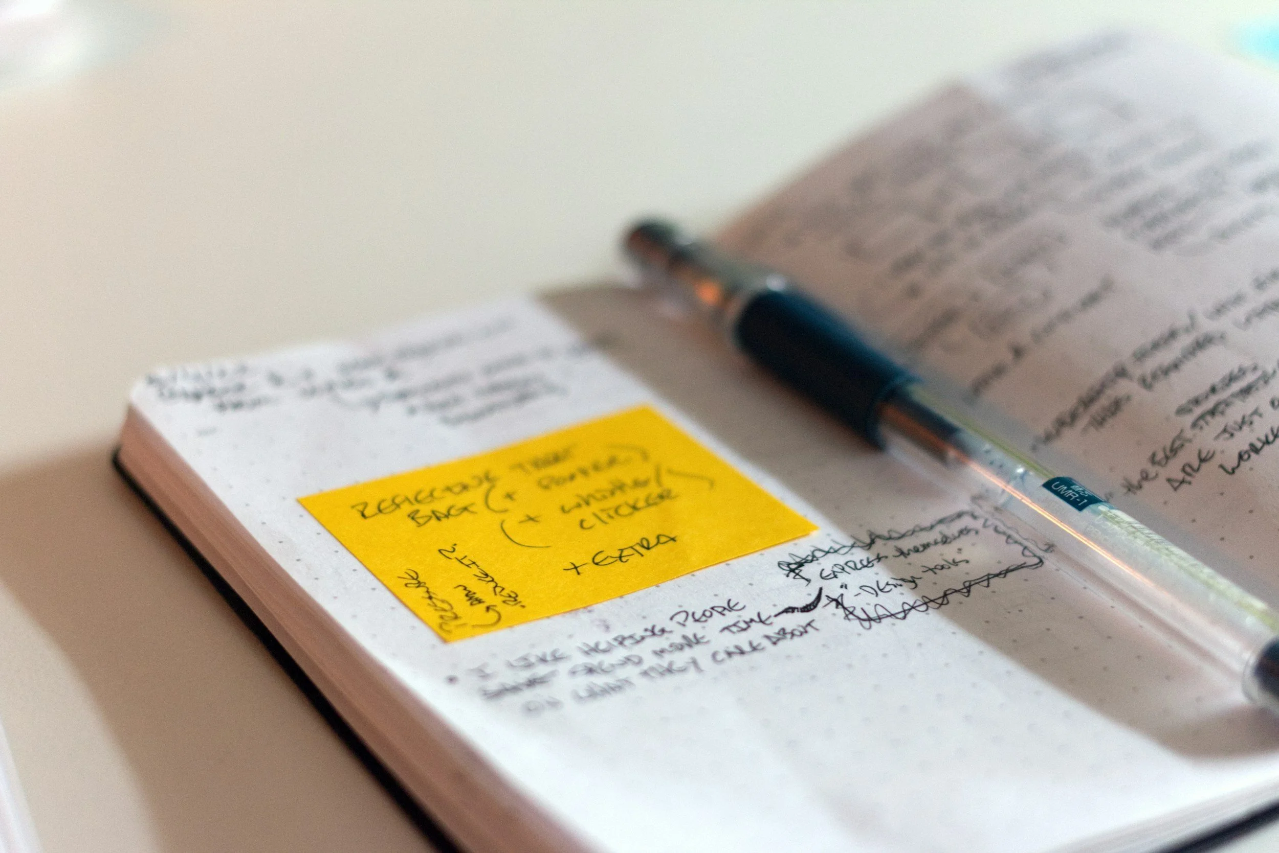 Open dotted notebook with handwritten notes, black pen resting on the page, and a yellow sticky note with handwritten text.