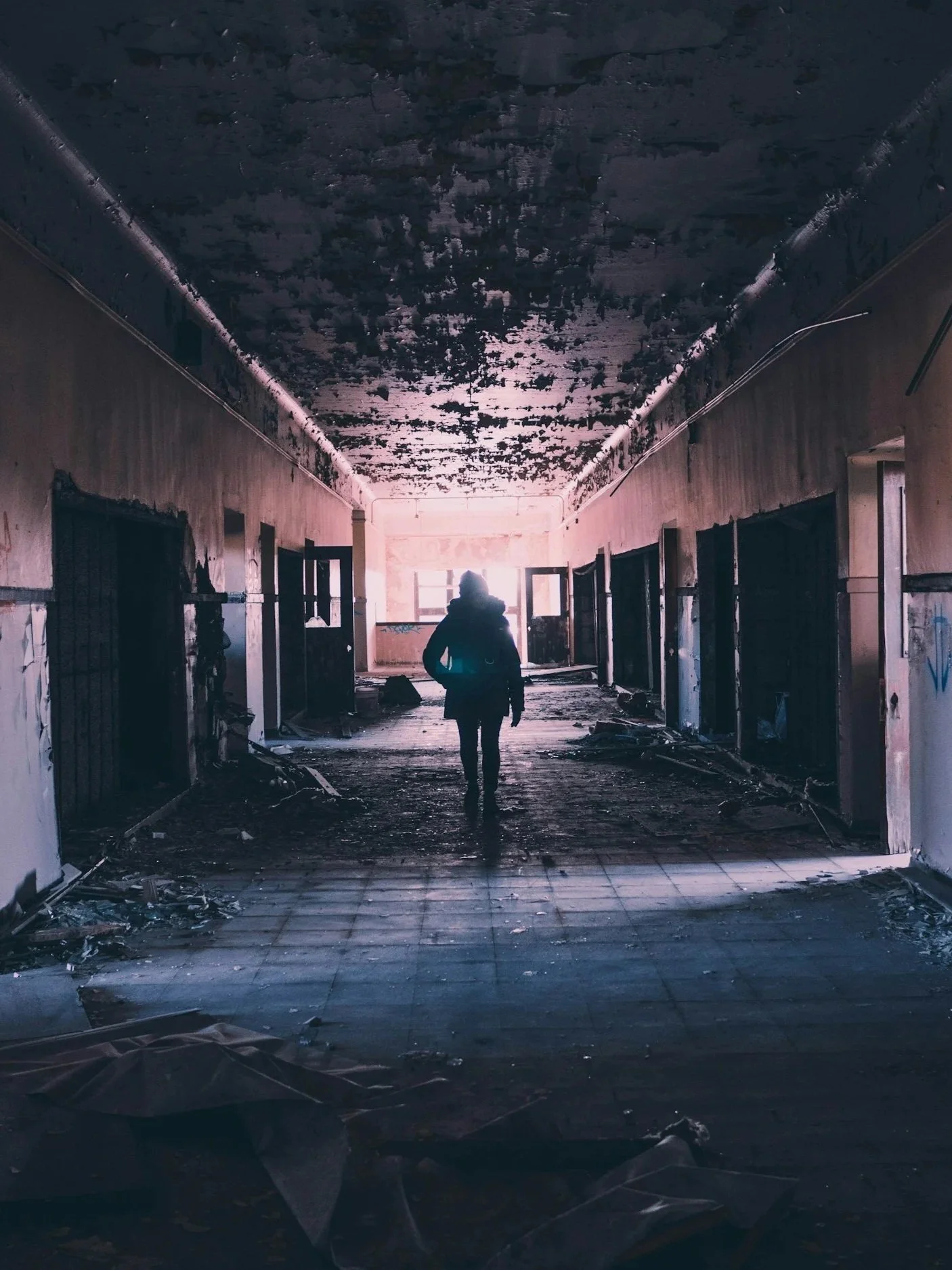 An abandoned and damaged hallway with charred ceiling and scattered debris, with a silhouette of a person walking towards a bright exit at the end of the corridor.