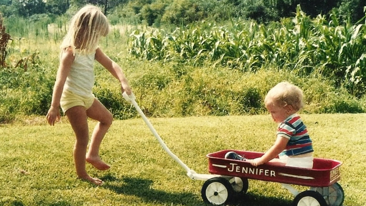 A young girl pulling a red wagon with a smiling toddler sitting inside, in a grassy field with corn in the background. The wagon has the name "Jennifer" written on its side.