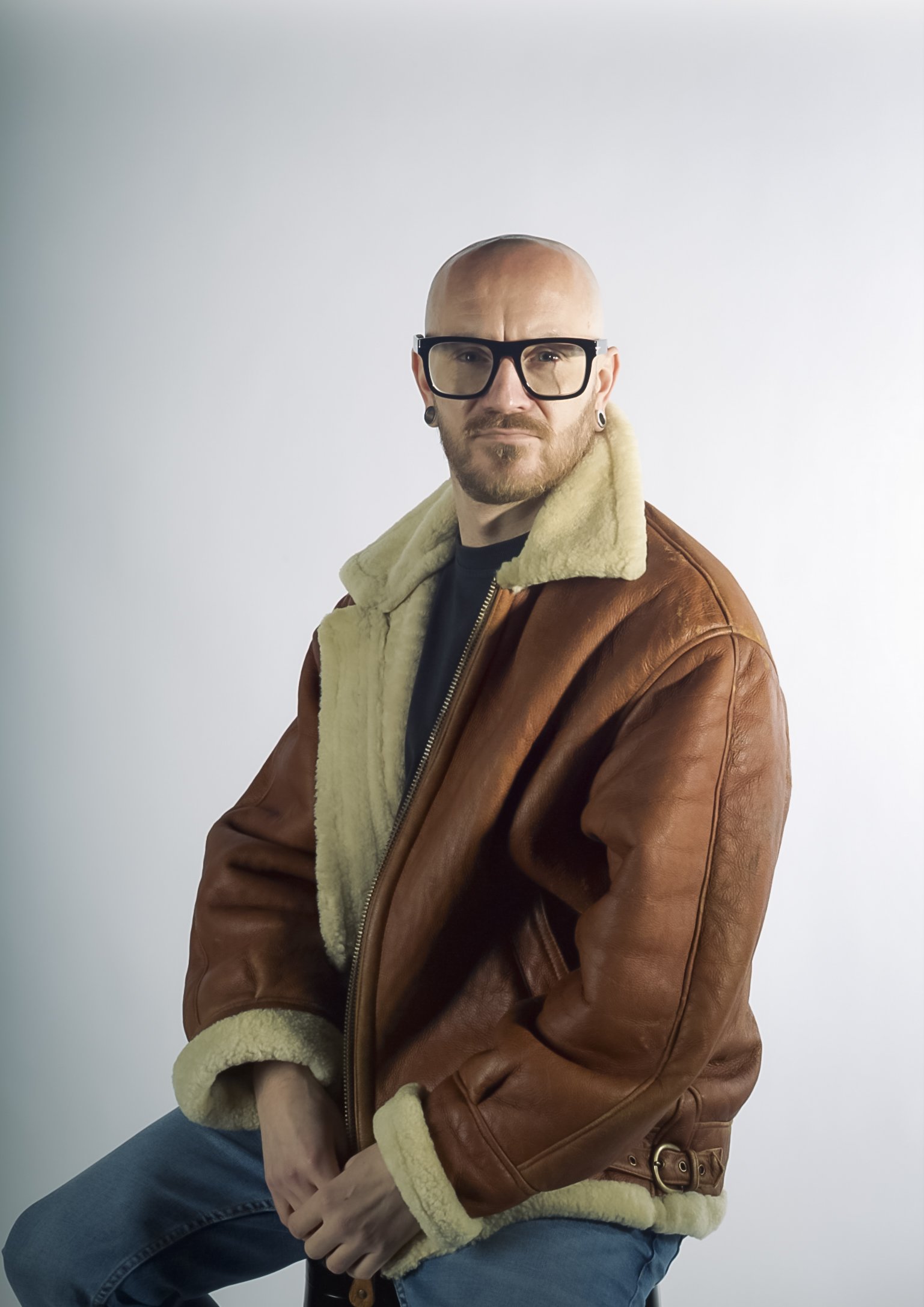 A man wearing black-rimmed glasses, a brown leather jacket with a cream-colored fleece lining, and blue jeans, sitting against a plain light background.