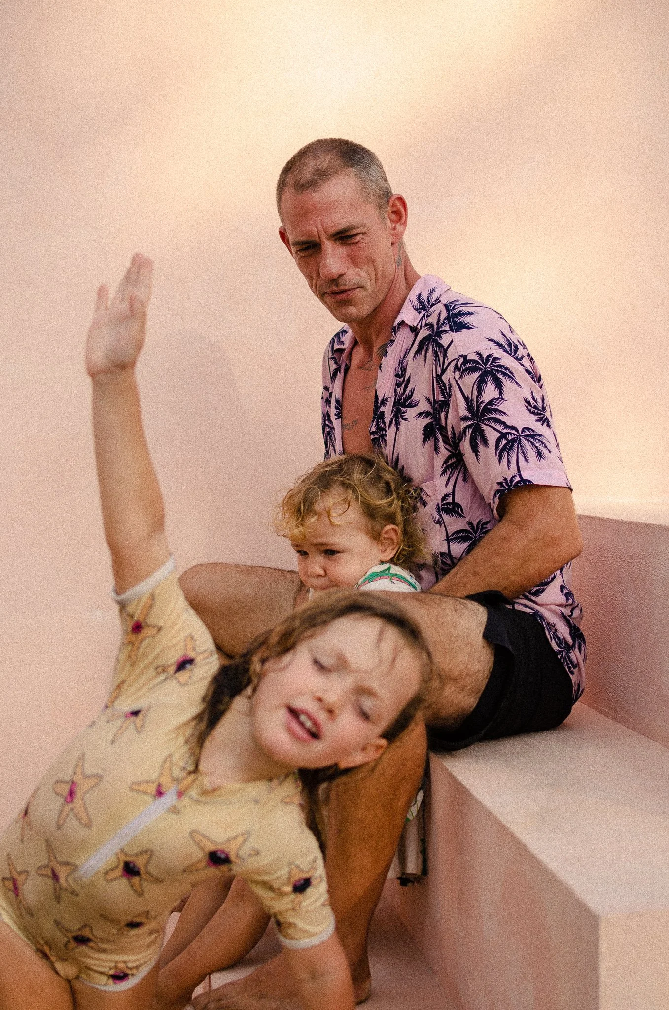 A man with short hair wearing a pink Hawaiian shirt with palm trees, sitting on pink steps, holding a young girl with curly blonde hair, wearing a colorful shirt, while another girl with curly hair in a beige star-patterned shirt leans against his le