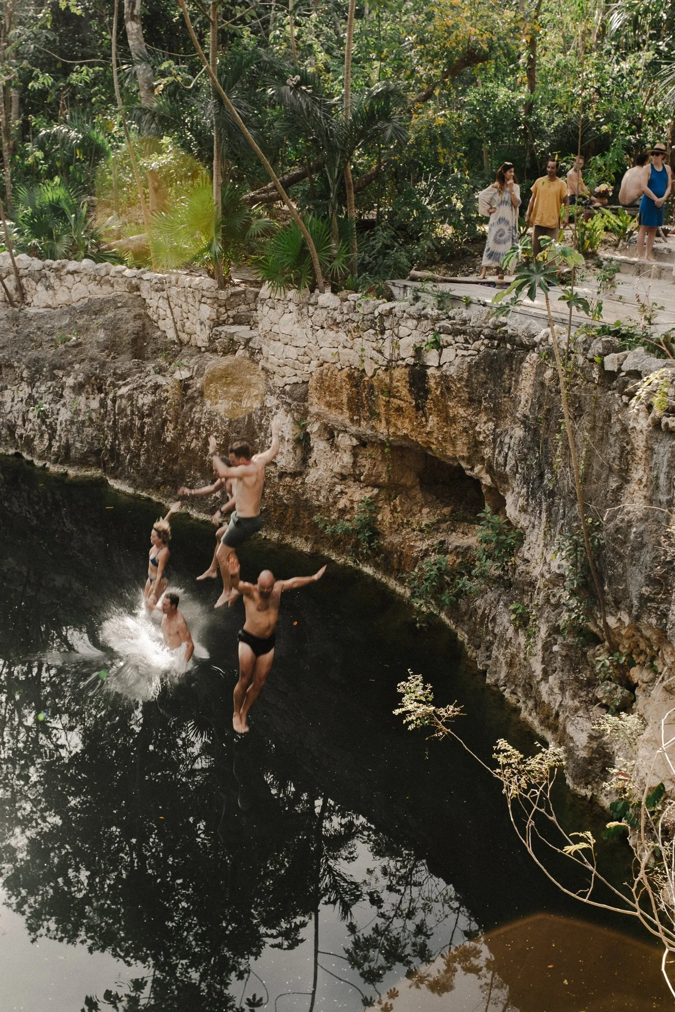 People jumping into a natural cenote with a rocky edge surrounded by lush greenery and trees.