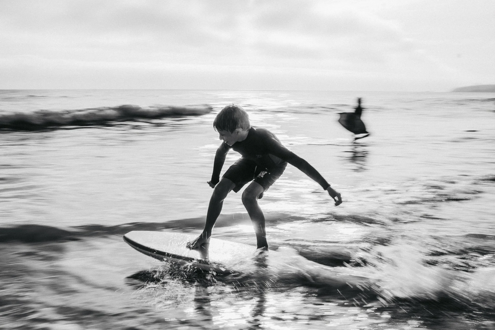 A boy surfing on the ocean with another surfer in the background during sunset, black and white photo.
