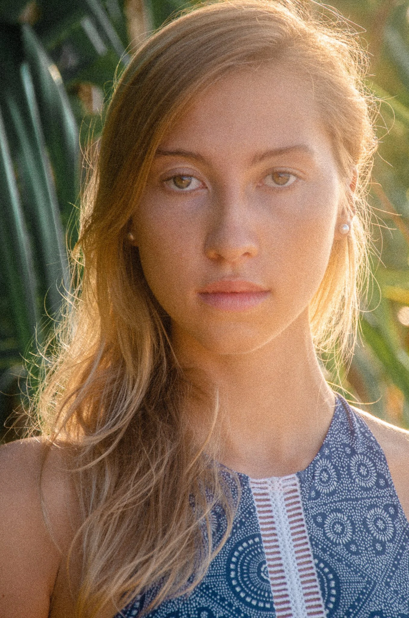 Close-up of a young woman with blonde hair, wearing a patterned blue sleeveless top and pearl earrings, outdoors with greenery in the background.