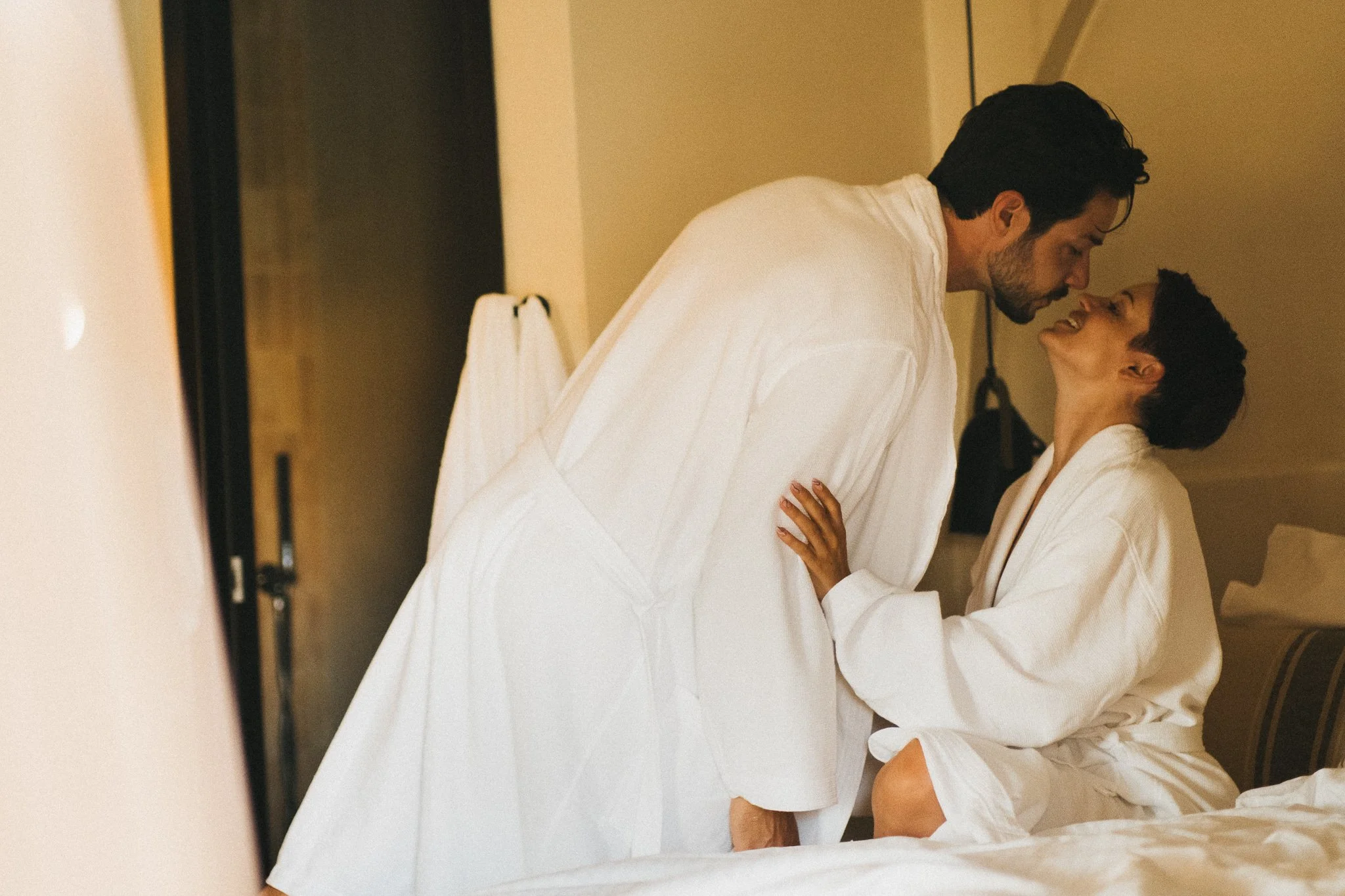 A couple in white robes sharing an intimate moment in a bedroom, with the woman sitting on the bed and the man leaning toward her.
