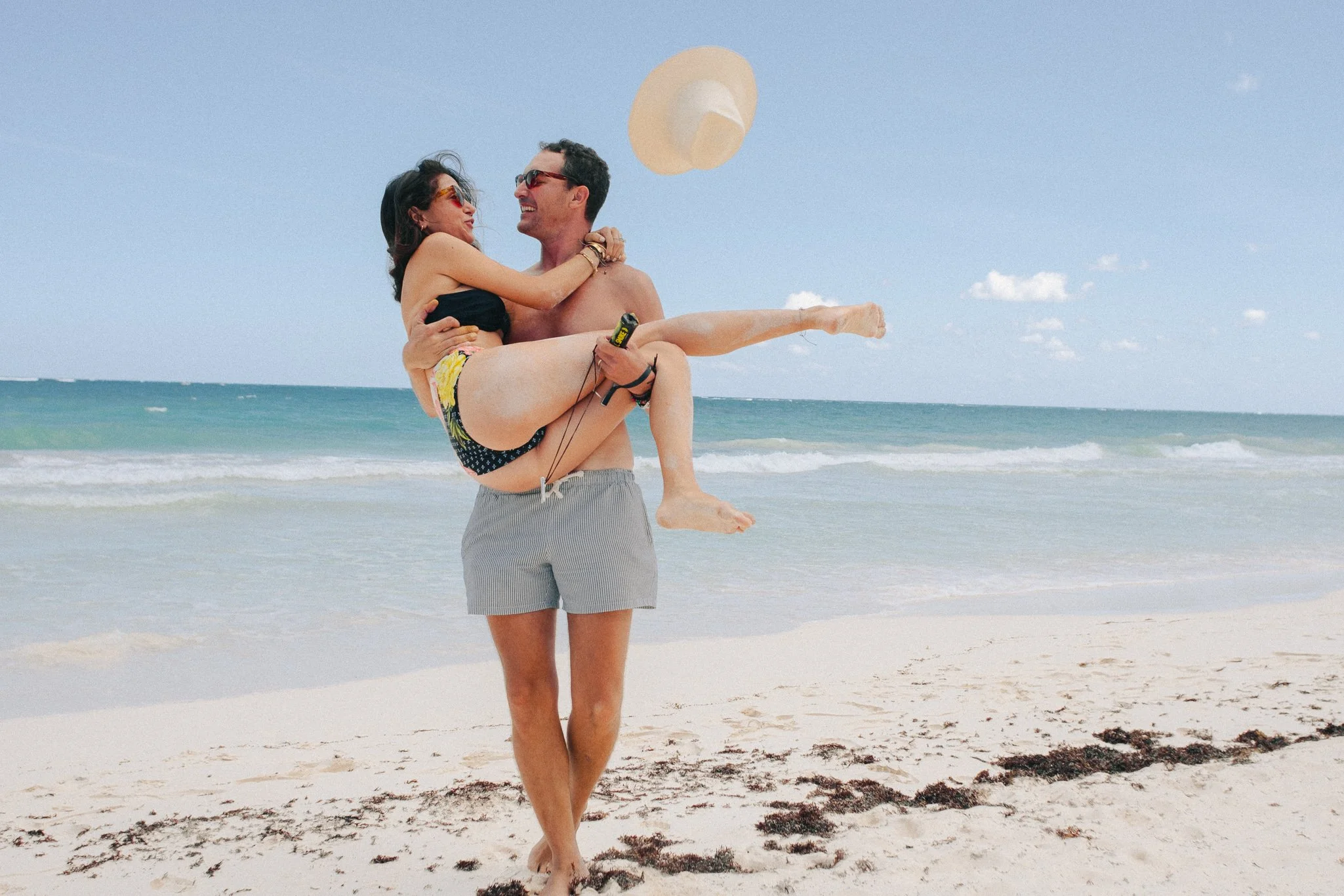 A man holding a woman in his arms on the beach, both smiling and looking at each other. The woman is wearing a black top and colorful shorts, the man is shirtless and wearing gray shorts. There is a large sun hat floating above them and the ocean in 