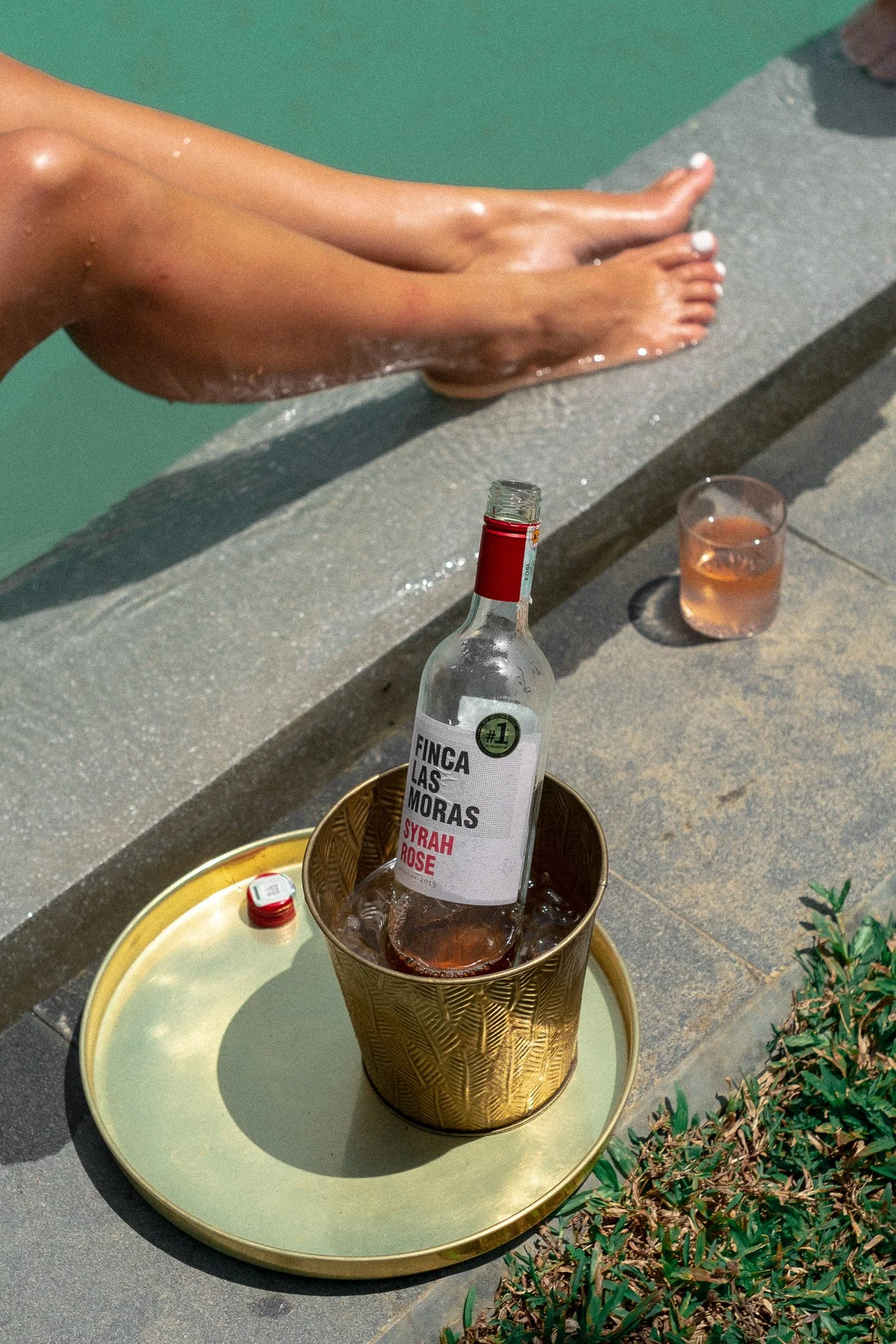A person sitting on the edge of a pool with their feet in the water, a bottle of wine in an ice bucket, and a glass of rosé on the poolside.