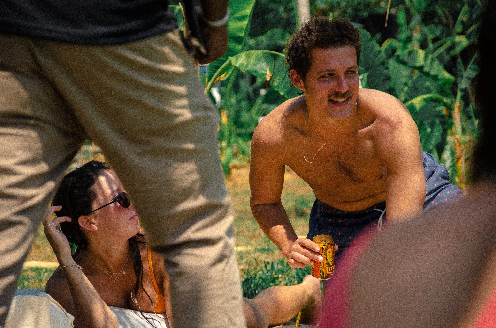 A shirtless man with a mustache and curly hair smiling while holding a can of beverage, sitting outdoors with green plants in the background. A woman wearing sunglasses and a spaghetti strap top is sitting on the ground nearby, partially obscured by another person's legs.