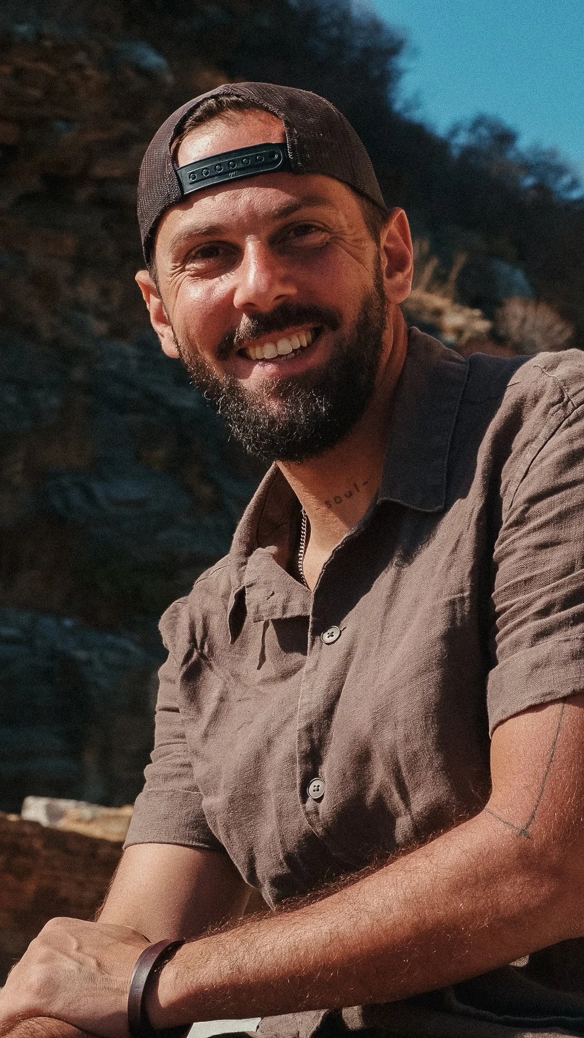 A smiling man with a beard wearing a backwards cap and a short-sleeved button-up shirt outdoors at dusk.