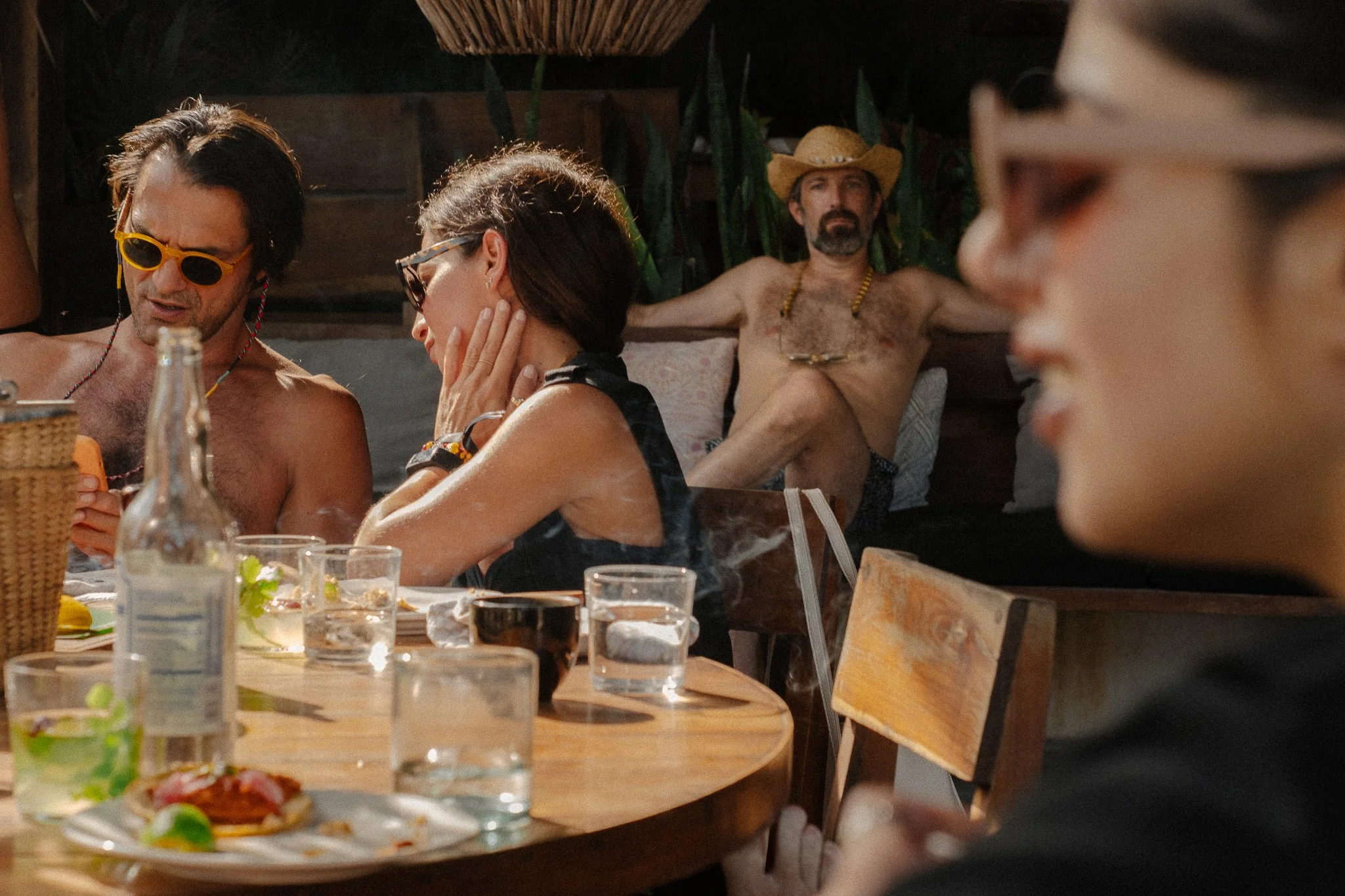 Group of four adults at an outdoor party, one man with yellow sunglasses talking to a woman, a man in a cowboy hat relaxing in the background, and a woman in glasses in profile, with a table of food and drinks in front of them.