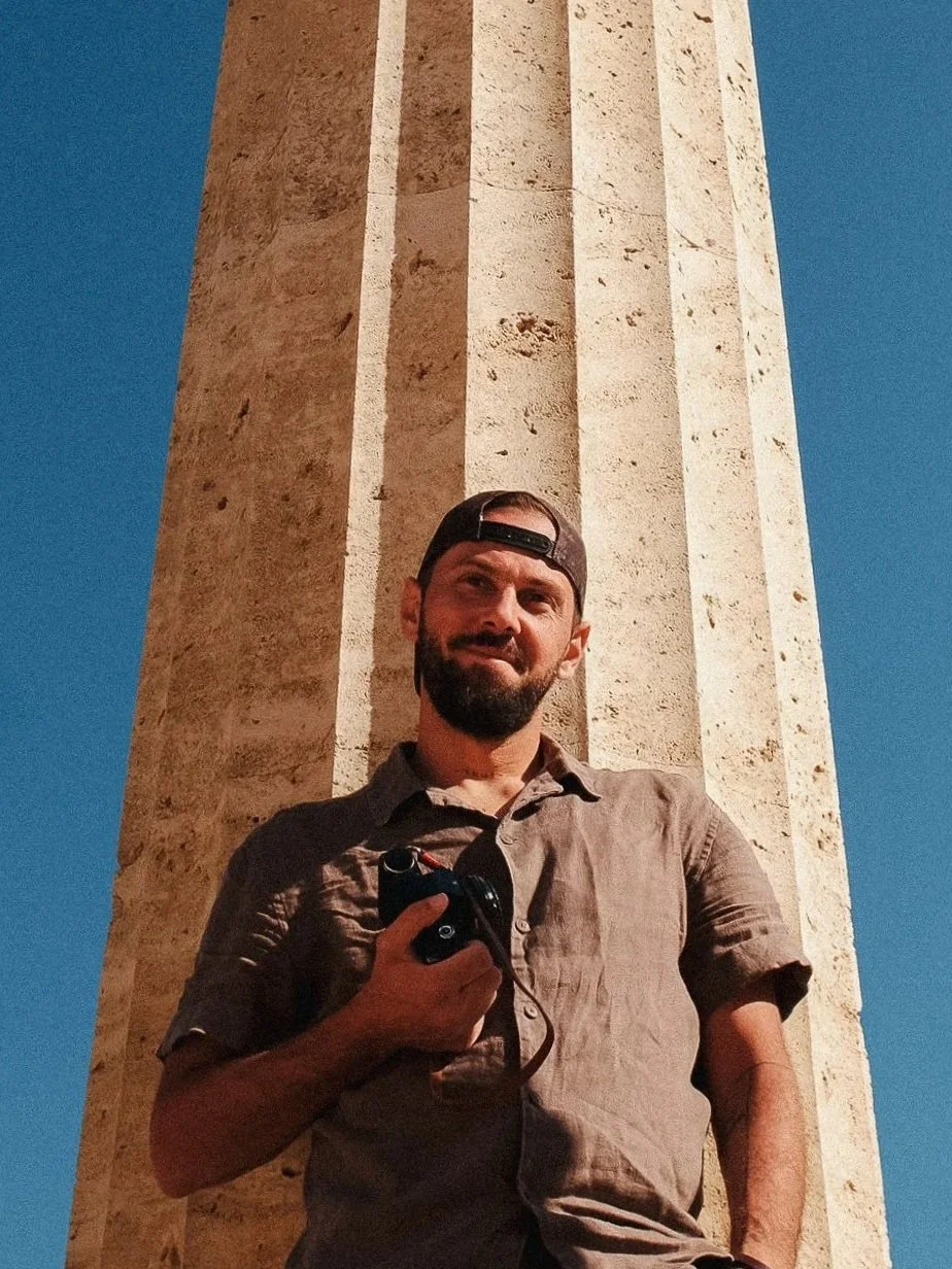 A man with a beard and cap holding a camera, standing in front of a large stone pillar with a clear blue sky in the background.