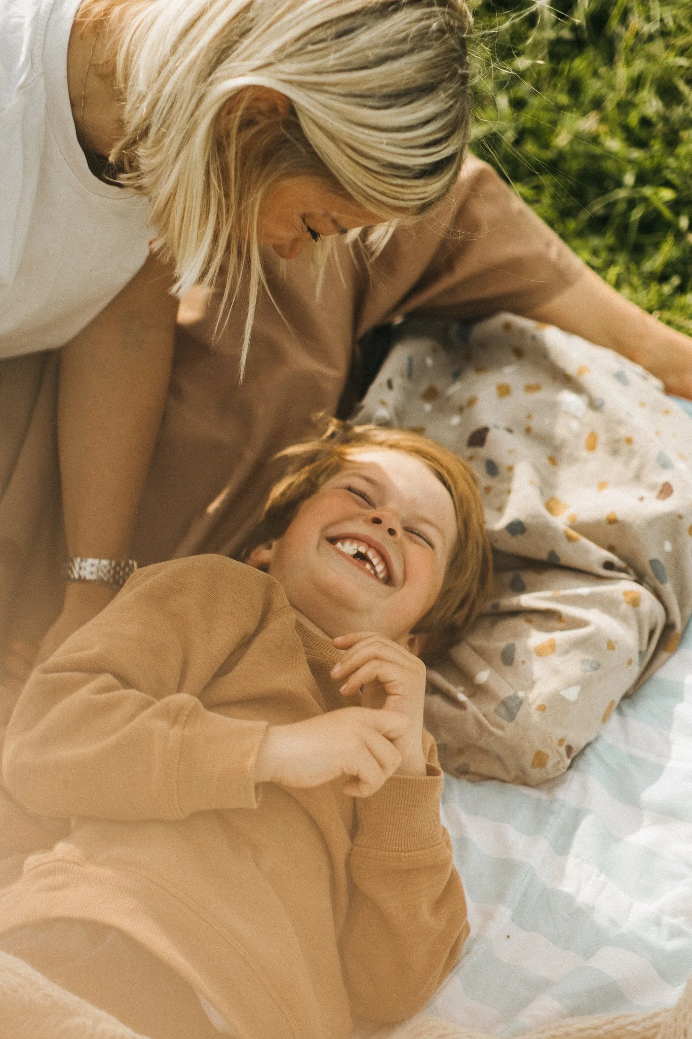 A young boy with red hair lying on a bed outdoors, smiling and laughing, with a woman leaning over him, on a blanket with a terrazzo pattern.