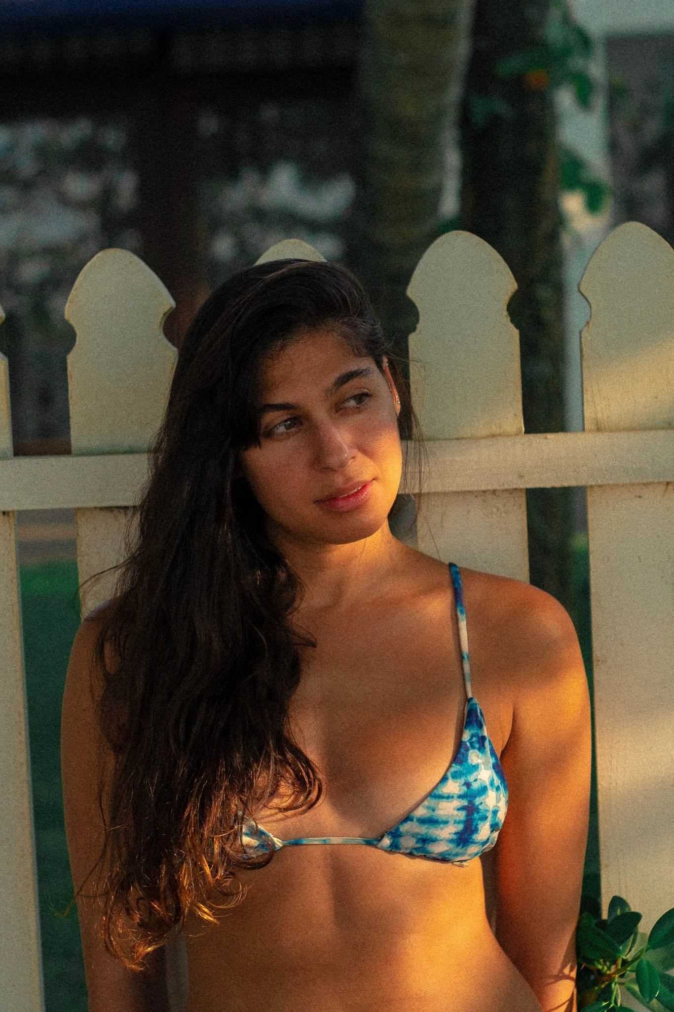 Young woman with long dark hair in a blue and white bikini top, standing next to a white picket fence, illuminated by warm sunlight during sunset.