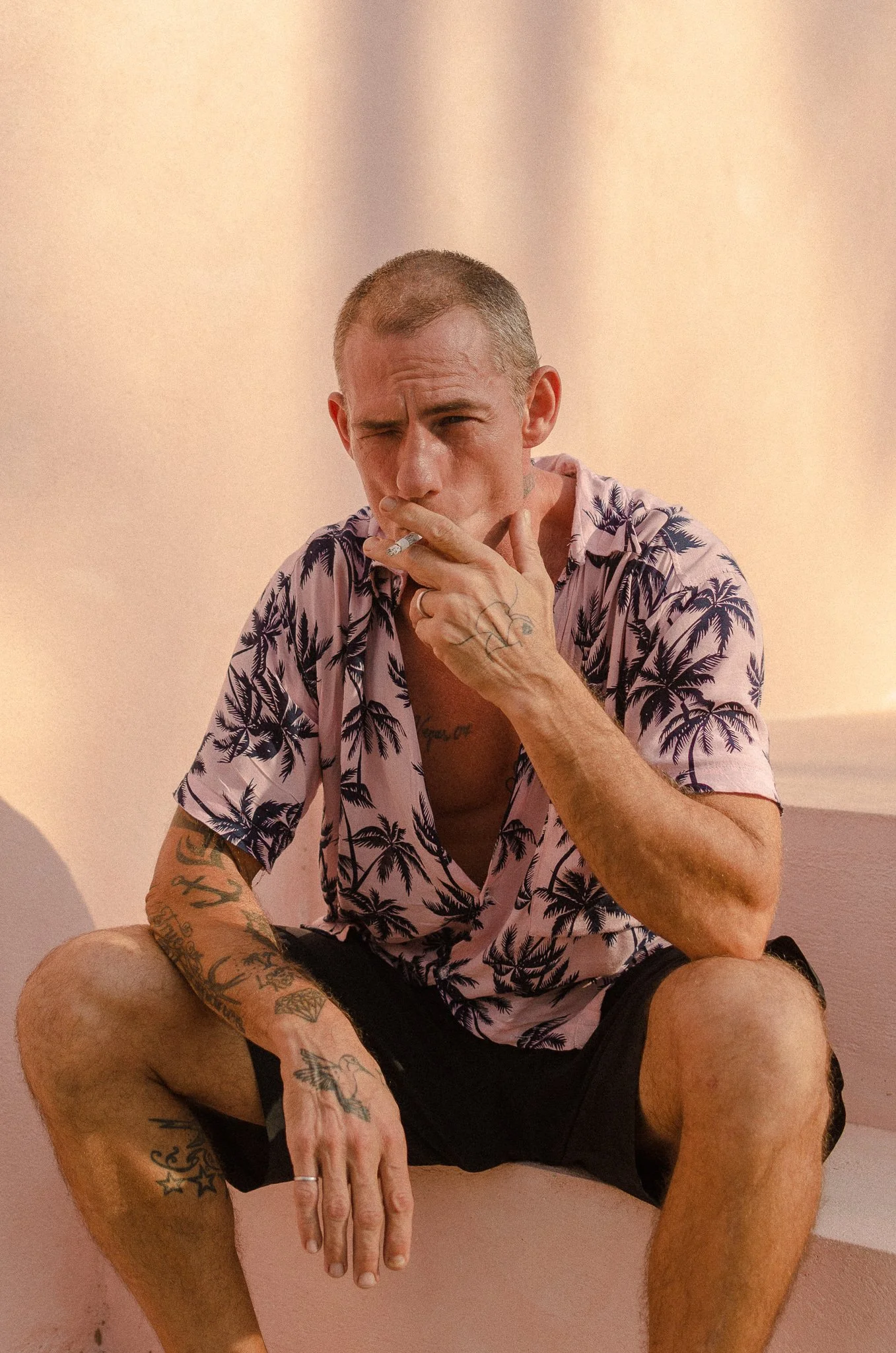 A man with tattoos and a short haircut, wearing a pink Hawaiian shirt with black palm trees, sitting on a step, smoking a cigarette.