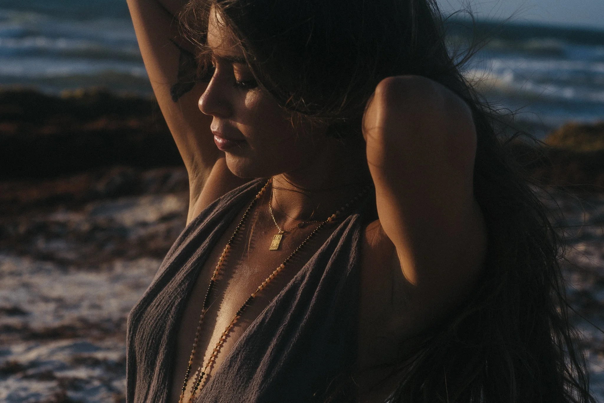 A woman with long dark hair standing on a beach during sunset, with her arms raised and eyes closed.