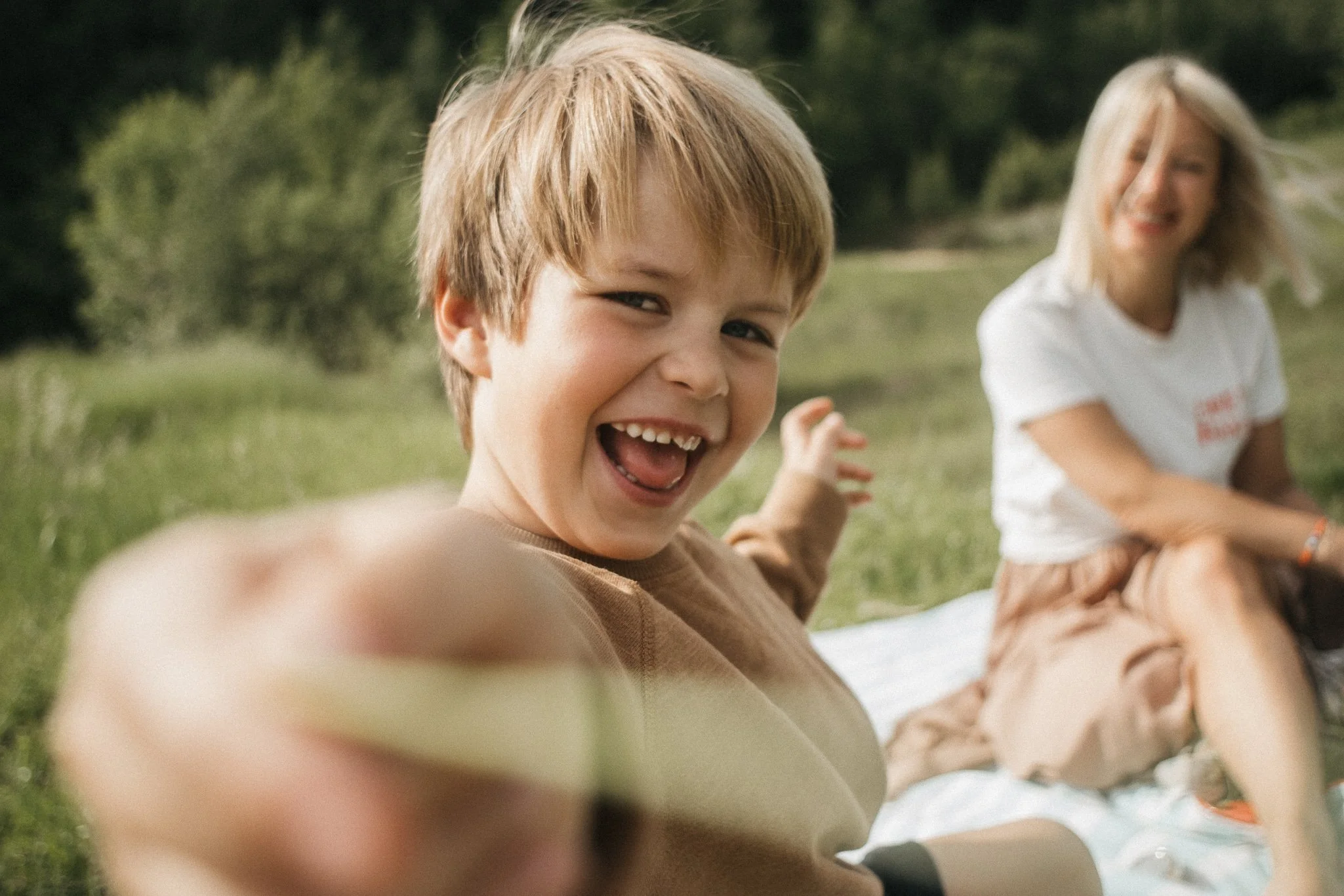 A young boy smiling and reaching towards the camera, with a woman sitting on a blanket in the background outdoors.