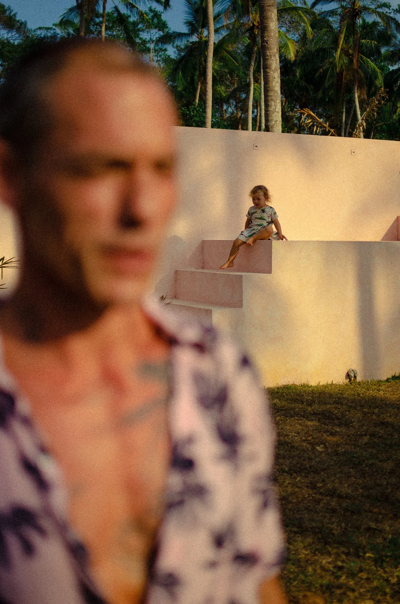 A blurred woman in the foreground with a child's face, and a young girl sitting on a staircase in the background among palm trees, during sunset.