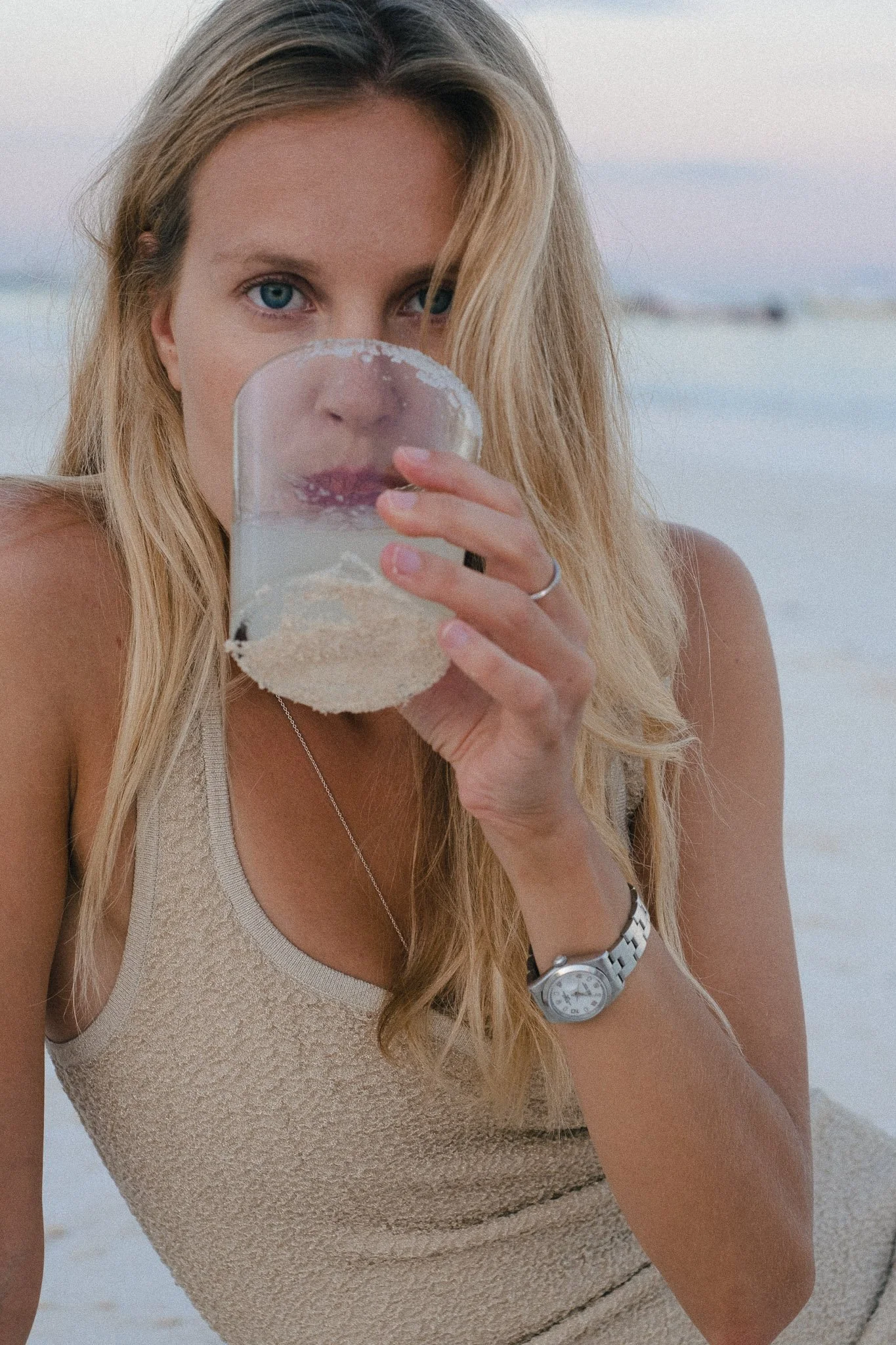 A woman with blonde hair and blue eyes at the beach, holding a transparent glass with sand and water, and drinking from it.
