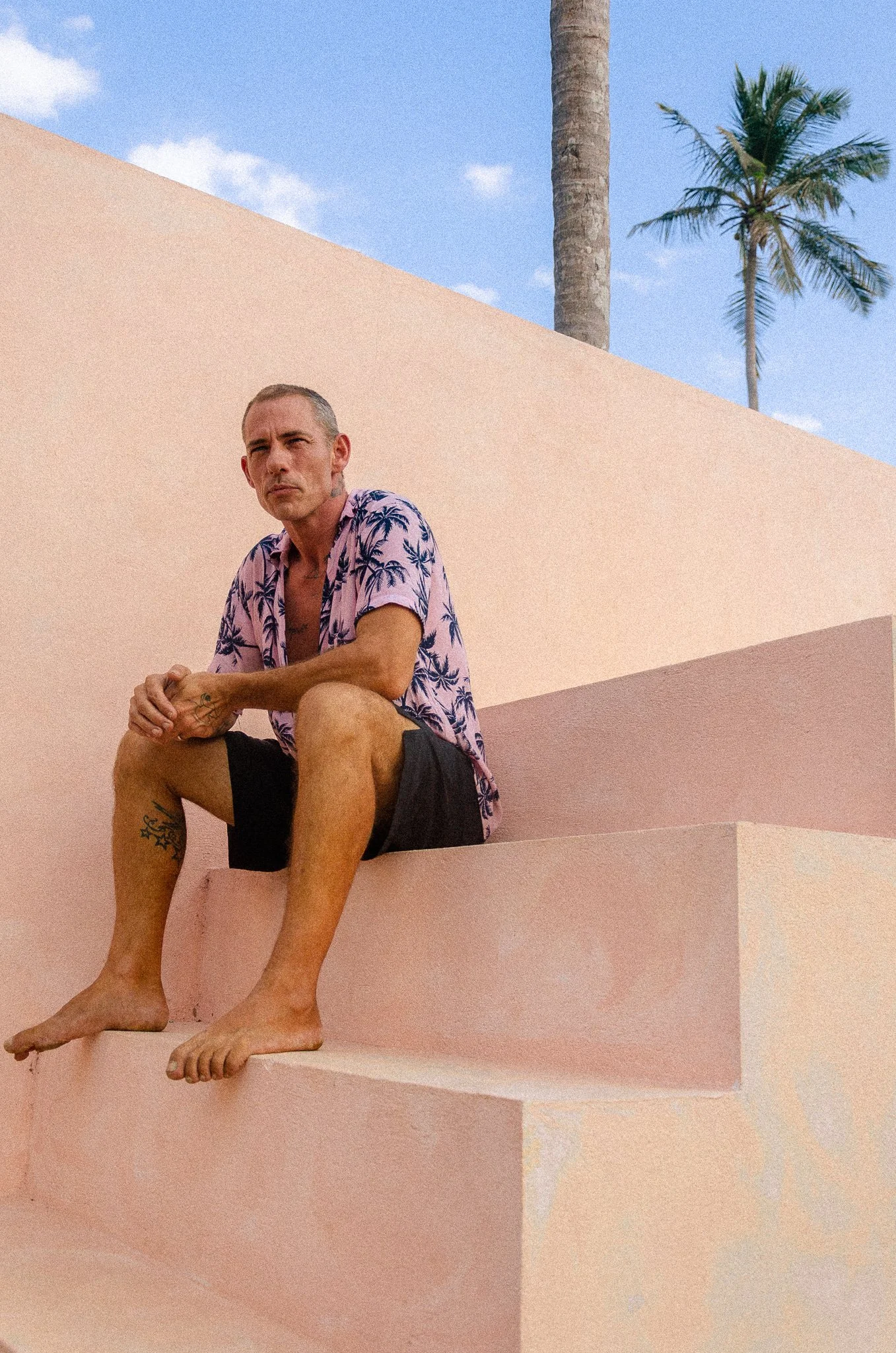 A man sitting on pink concrete steps outdoors with palm trees and a blue sky in the background.