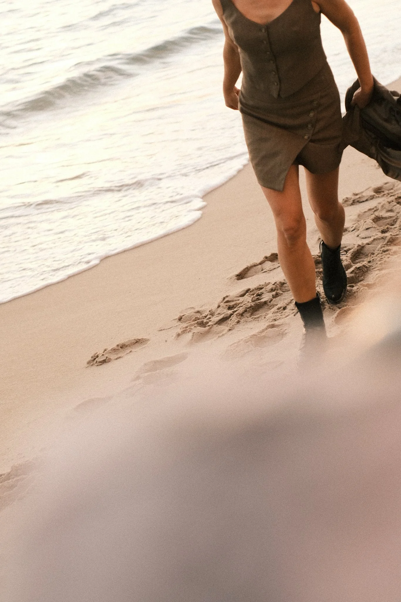 Person walking on the beach in a brown dress, wearing black shoes and carrying a backpack, near the water's edge.