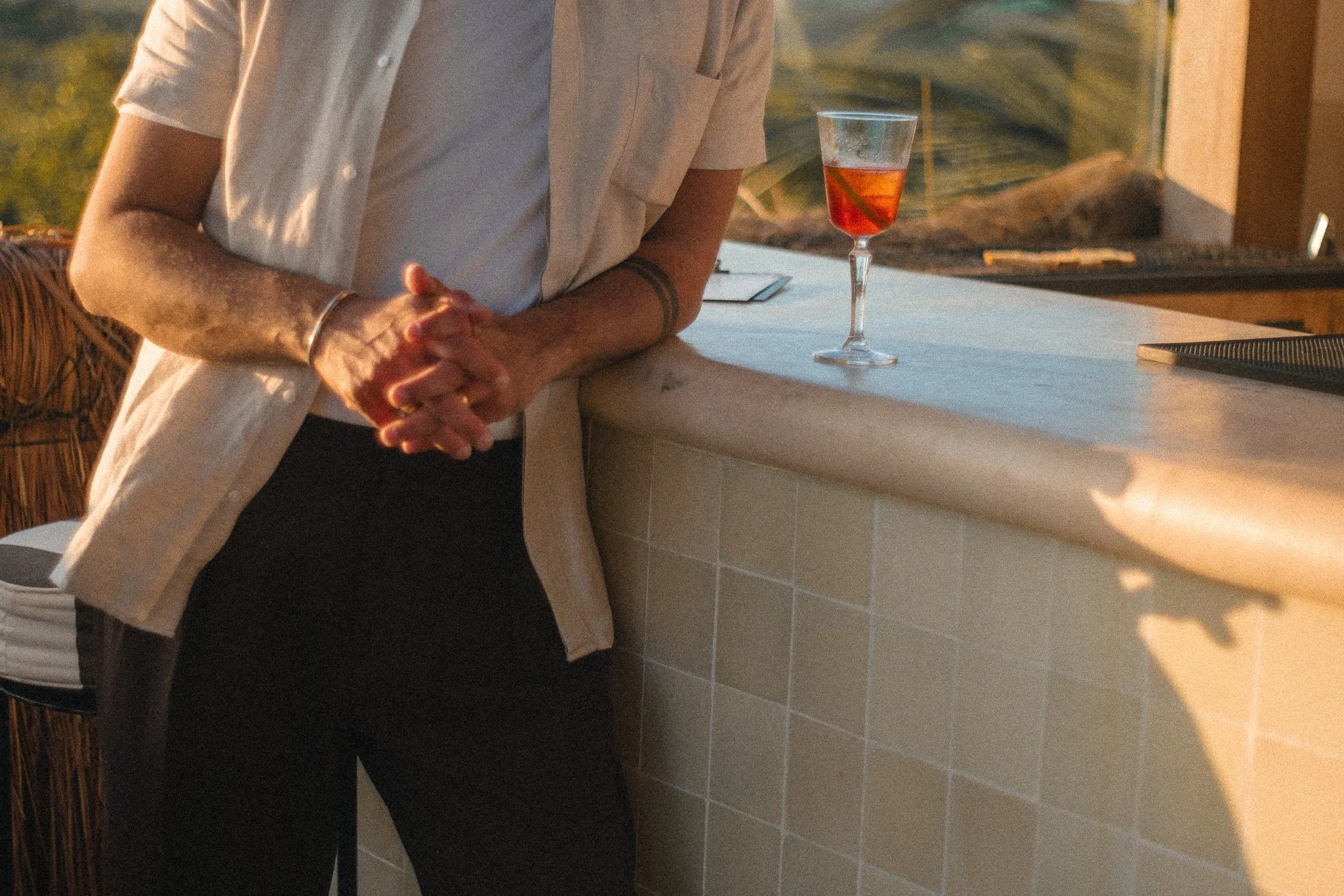 Person standing at a bar counter with their hands clasped. There is a glass of red cocktail or wine on the counter. The setting appears to be during sunset or in warm lighting.