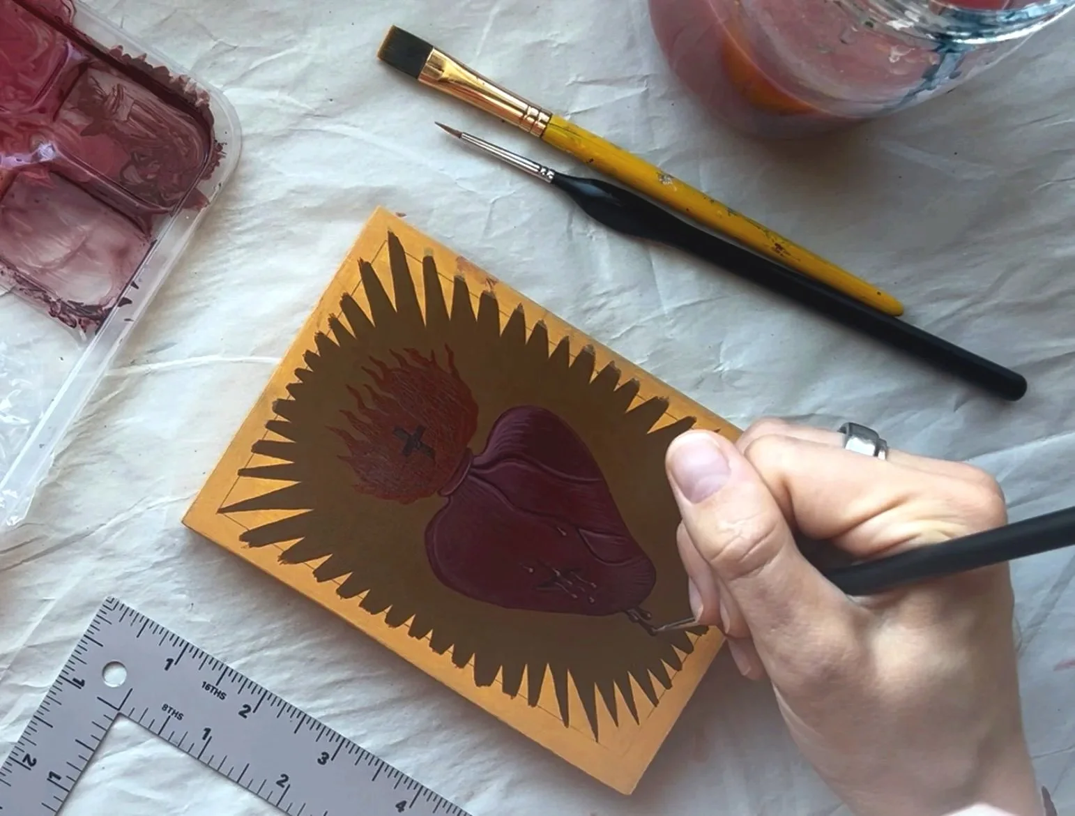 A person is creating a religious icon painting of the Sacred Heart of Jesus on a small wooden panel, using brushes and paints. The panel features an image of a heart with flames and a crown of thorns, surrounded by a golden background. Art supplies s