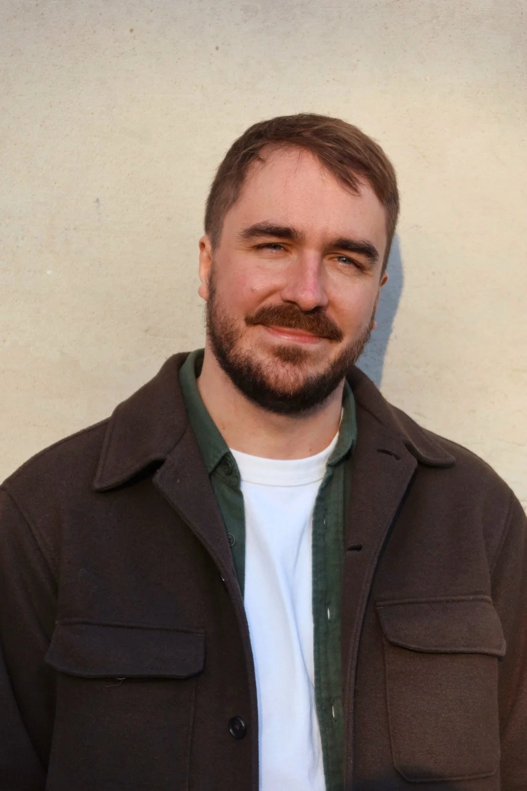 Un homme avec une barbe et des cheveux bruns courts, portant une veste foncée, une chemise verte et un T-shirt blanc, debout contre un mur beige.