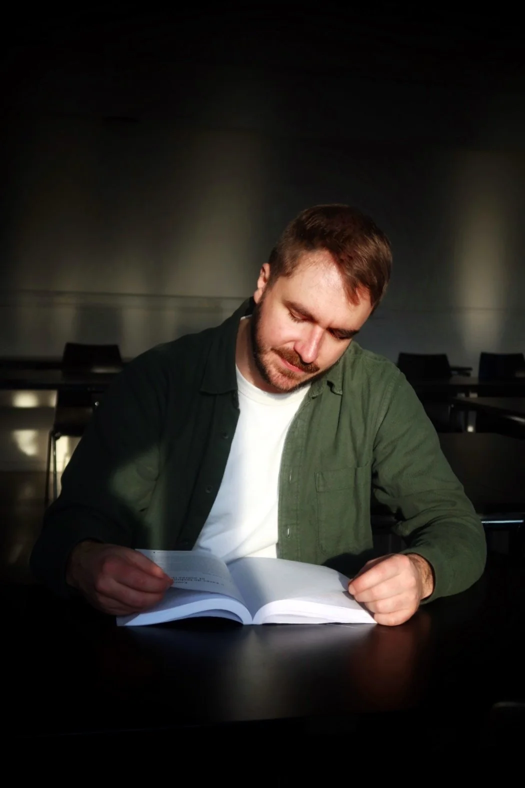 Un homme assis à une table, lisant un livre, dans une pièce sombre avec une lumière concentrée sur lui.
