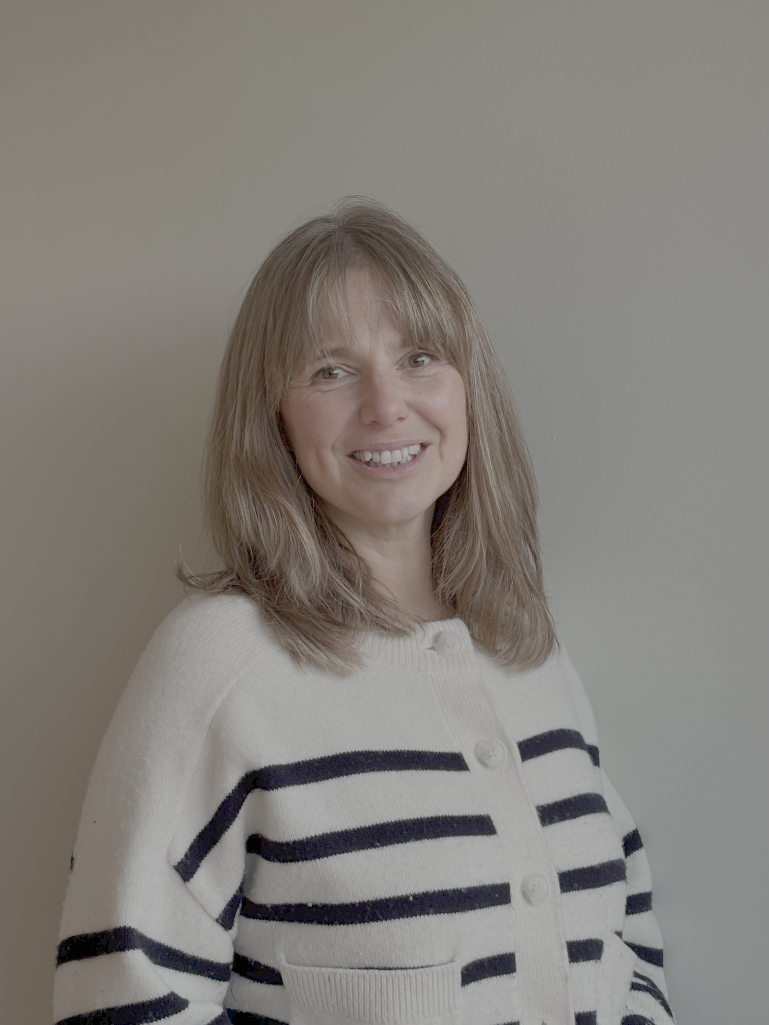 A woman with shoulder-length light brown hair, smiling, wearing a white cardigan with black horizontal stripes, standing against a plain beige wall.