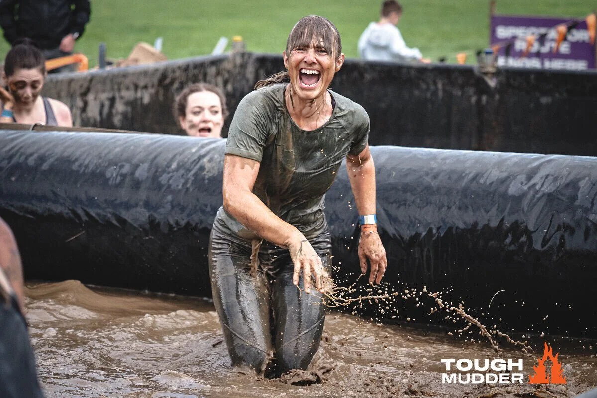 Woman smiling and muddy, emerging from water during a Tough Mudder obstacle course event, with other participants in the background.