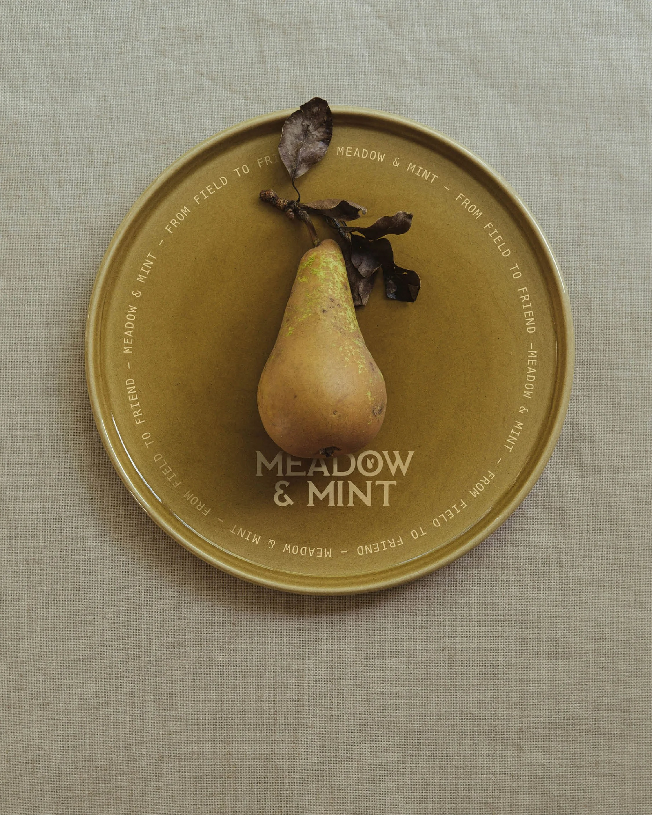 A pear with dried leaves on a green plate with the words 'Meadow & Mint' and a decorative circular text, placed on a textured beige surface.