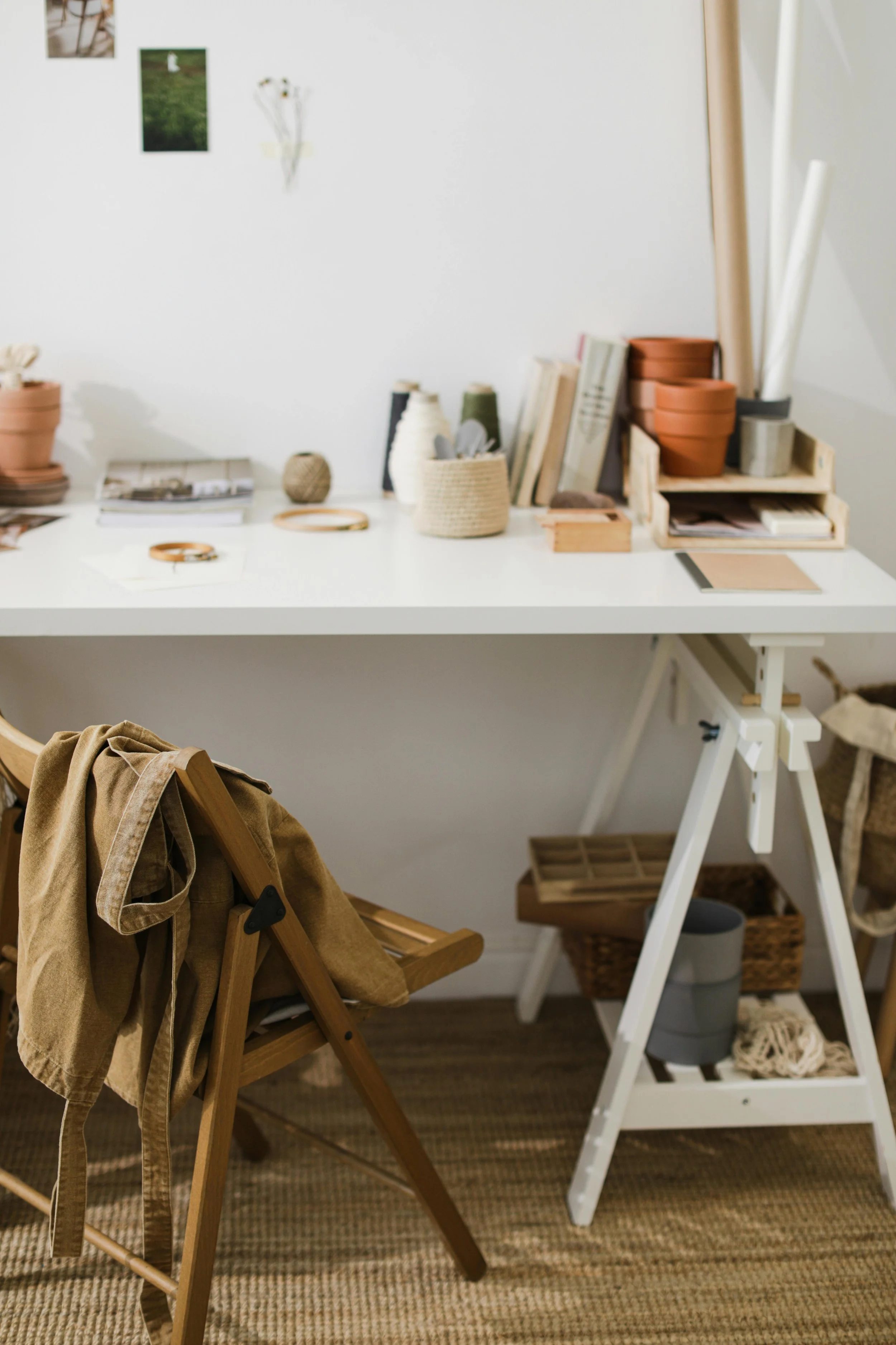 A white desk with various potted plants, books, and decorative items on top. A wooden chair with a tan jacket draped over it stands beside a white adjustable table, with baskets and storage underneath.