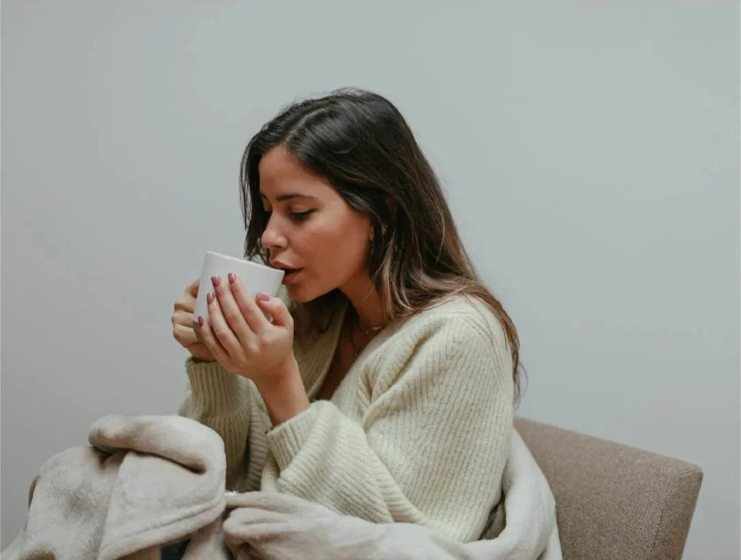 Une femme brune avec des cheveux ondulés, portant un pull beige, profite d'une pause avec une tasse blanche, assise dans un salon discret avec un fond blanc