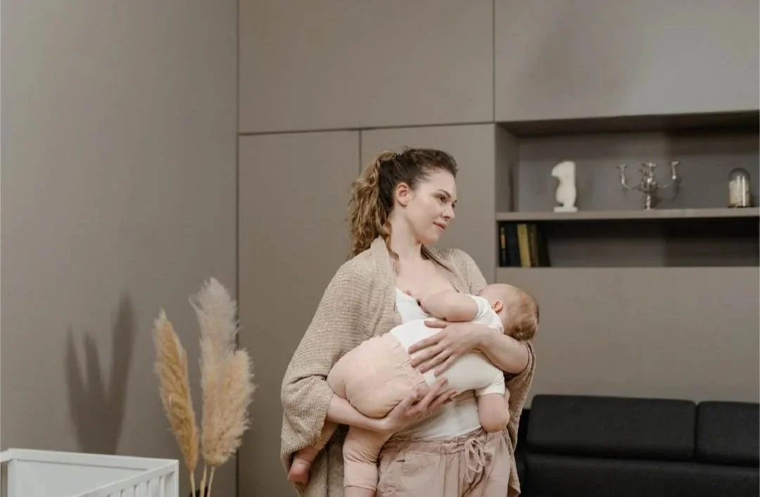 Une femme tient un bébé dans ses bras, dans un intérieur moderne, face à un fond gris avec des étagères et des décorations.
