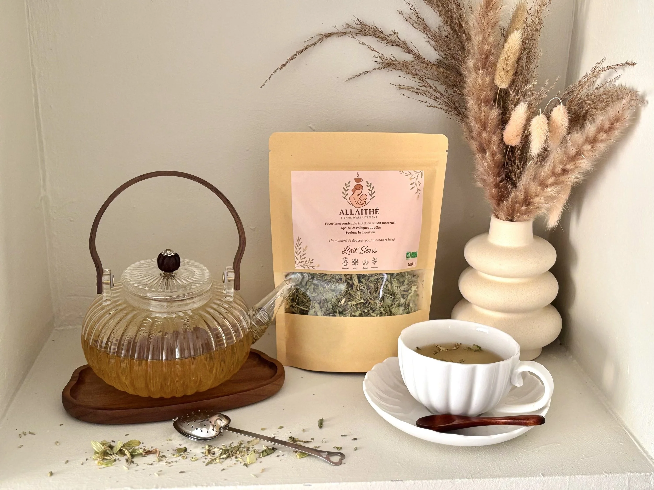 Une théière en verre avec du thé, un sac de tisane allaithe, une tasse de tisane accompagnée d'une cuillère en bois, une vase blanche avec des dried grasses et des fleurs séchées.