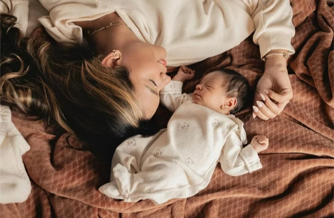 Une femme allongée sur un lit, yeux fermés, près d'un bébé qui dort.