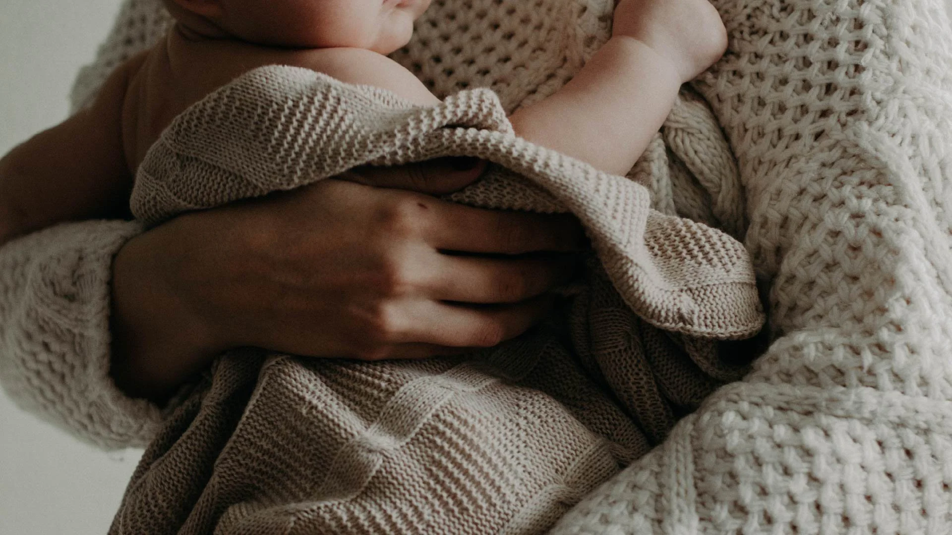 Une personne tenant un bébé contre elle, entourés de couvertures et vêtements en tricot de couleur beige et marron.