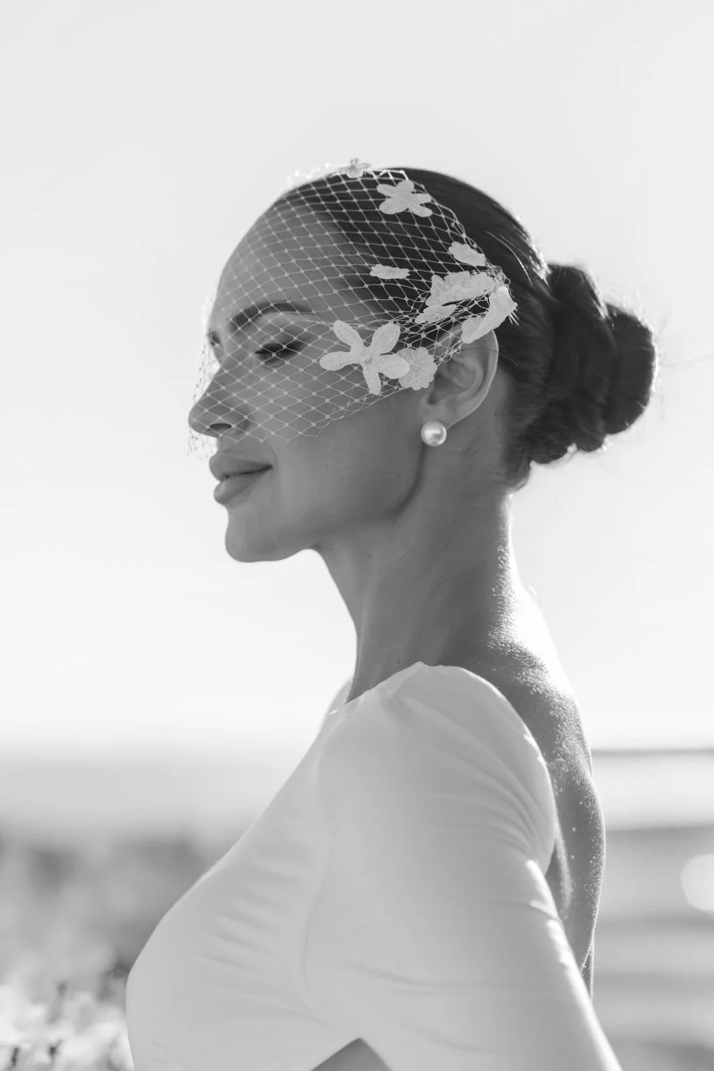 Black and white photo of a woman in profile, wearing a pearl earring and a lace veil with floral patterns, smiling gently.