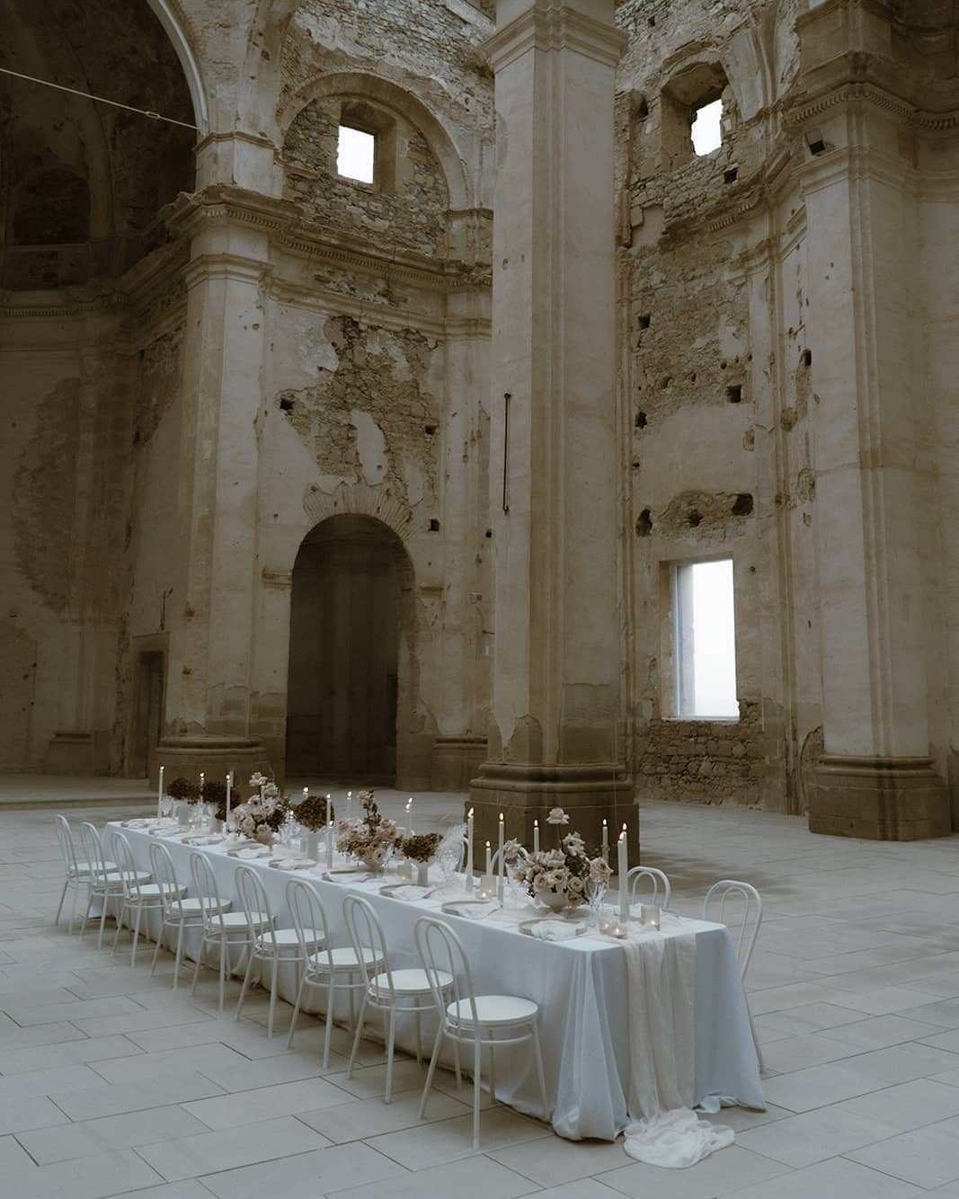 Elegant wedding reception setup with a long white table, floral centerpieces, and candles inside a historic, partially ruined stone building with high ceilings and large windows.