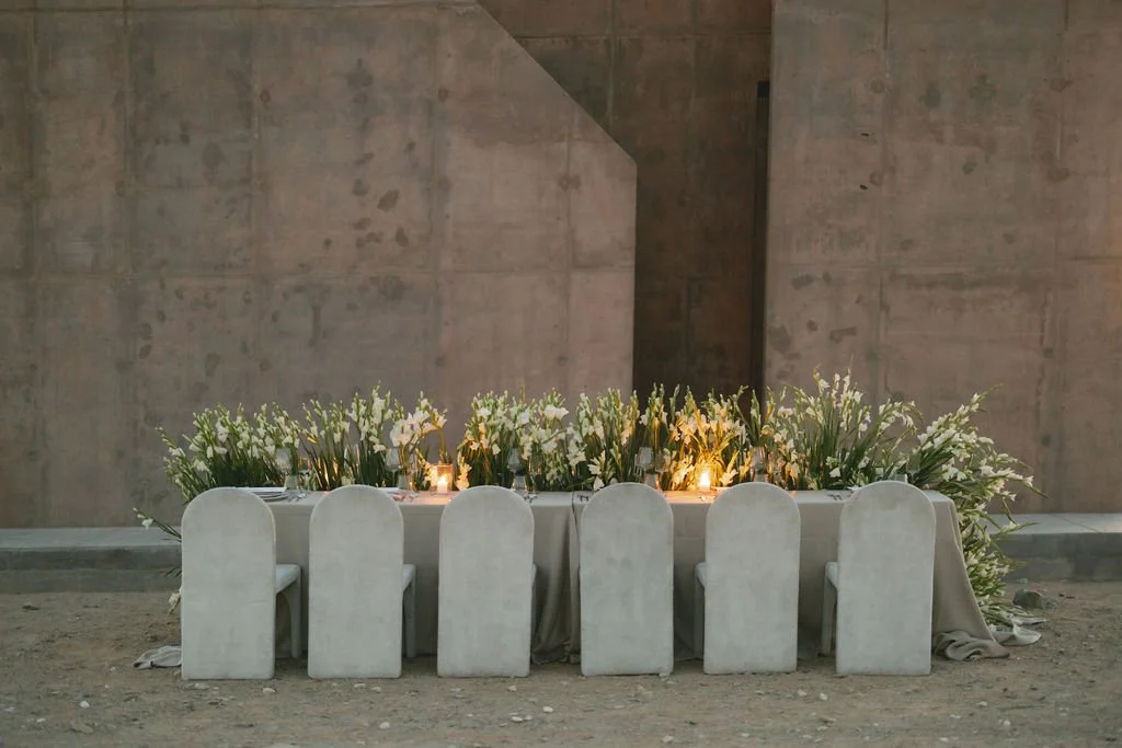 Elegant banquet table decorated with white flowers and candles, set against a modern cement wall.