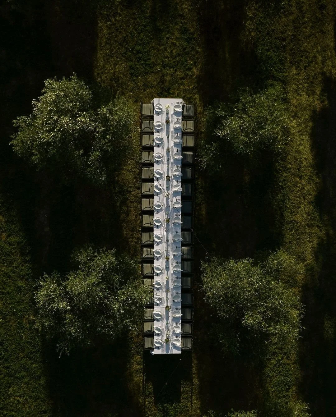 Drone view of long white outdoor event tent with tables and chairs, surrounded by green trees and grass.