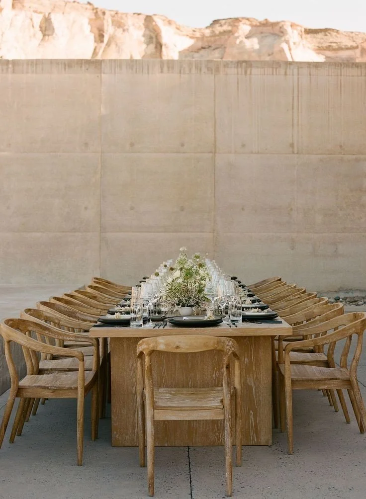 A long wooden dining table set with wine glasses, black plates, and a central floral arrangement of white flowers, surrounded by wooden chairs, in front of a tall concrete wall with rock formations in the background.