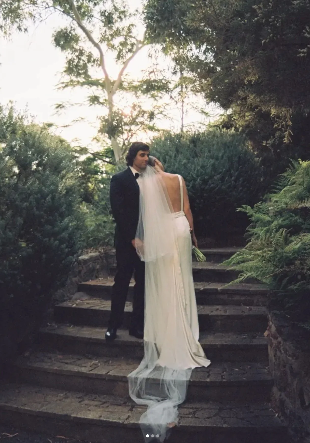 A bride in a white wedding dress and veil, and a groom in a black tuxedo, standing together on stone steps surrounded by lush greenery, outdoors during sunset.