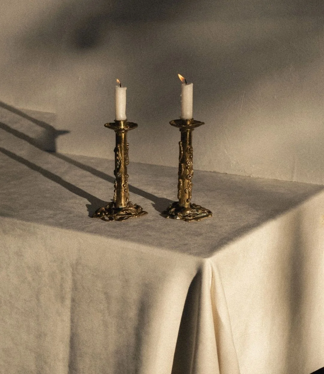 Two antique gold candlesticks with burning white candles on a neutral-colored cloth-covered surface, casting shadows on the wall.