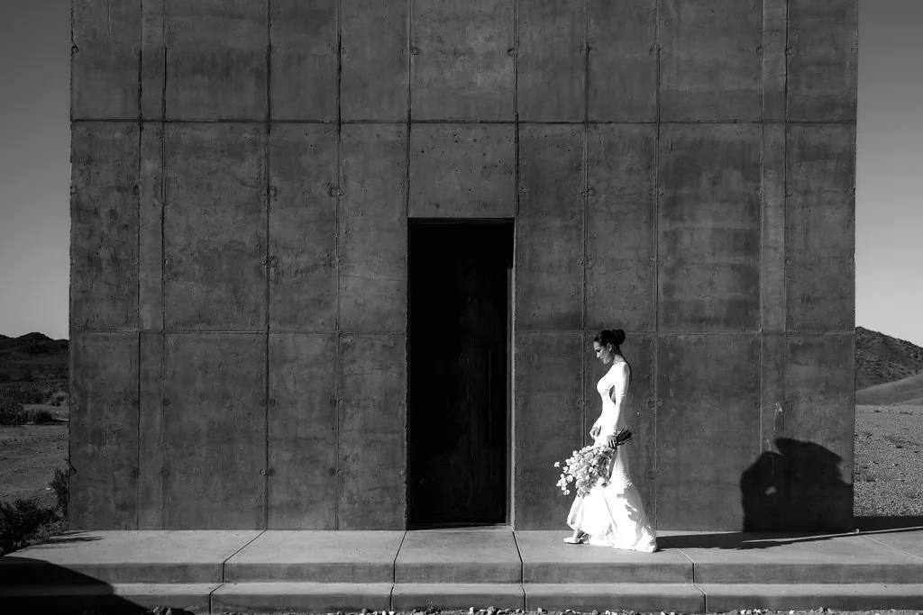 A woman in a white wedding dress holding a bouquet of flowers, standing outside a modern concrete building with a minimalist design, in a desert landscape.