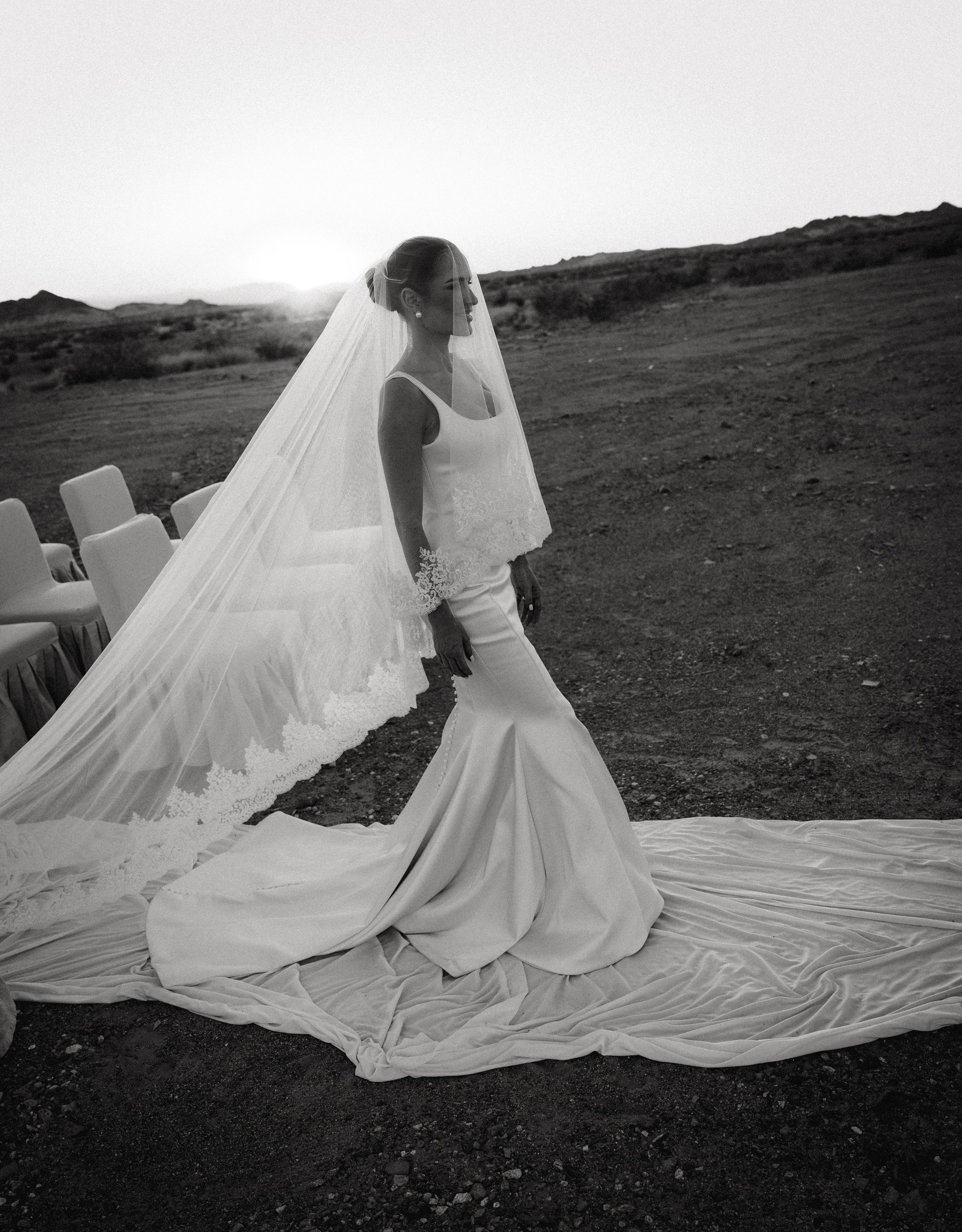 A bride in a wedding dress and veil stands on a rocky outdoor landscape with chairs set up in the background, capturing a black-and-white photo at sunset or sunrise.